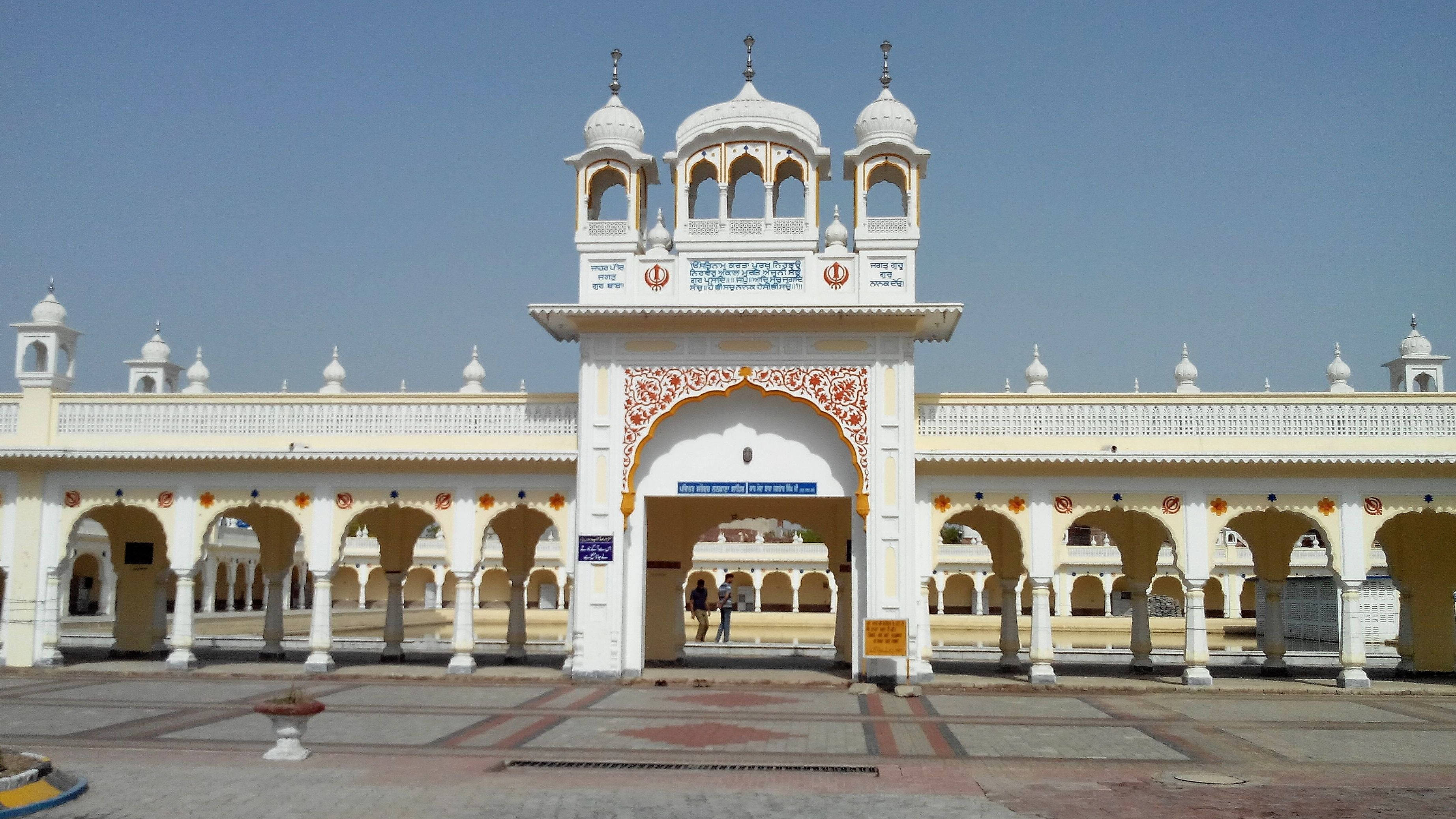 Lahore 桌布 壁紙 — 一座宏偉的白色錫克教謁師所，擁有華麗的拱門和圓頂，矗立在清澈的藍天下，有兩個人正走過入口。