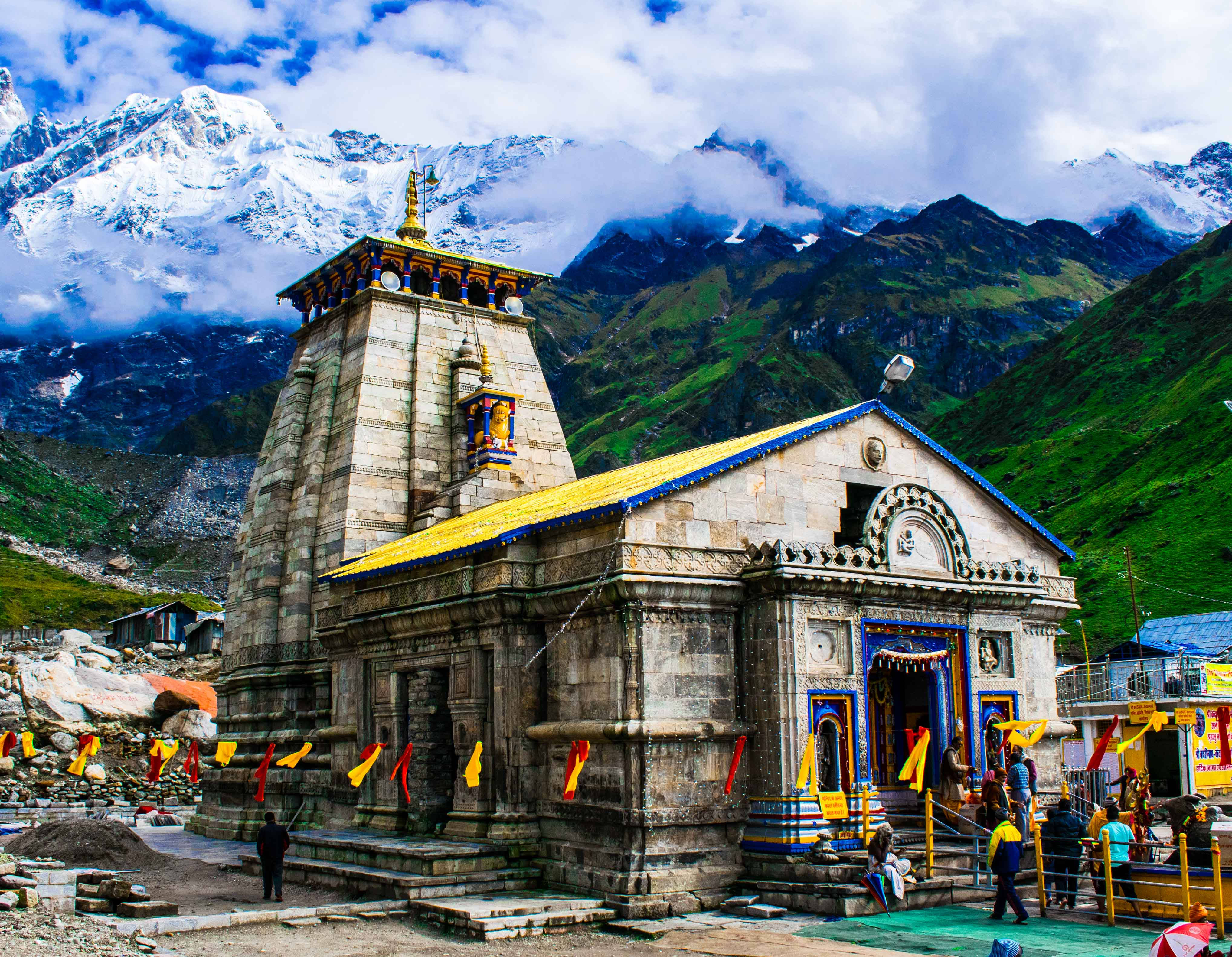 Kedarnath 桌布 壁紙 — 一座宏偉的石廟，金色屋頂，矗立在鬱鬱蔥蔥的青山和白雪皚皚的山峰之間，天空多雲。