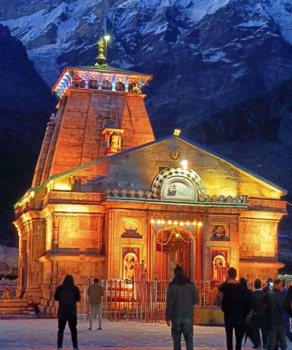 Kedarnath 桌布 壁紙 — 一座宏偉的石廟在暮色天空下，對著積雪的山峰溫暖地發光，遊客聚集在其燈火通明的入口前。