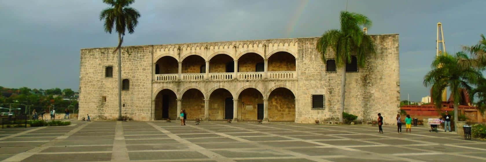 Architecture 桌布 壁紙 — 一座宏偉的石建築，擁有拱形走廊和陽台，矗立在多雲的天空下，一道淡淡的彩虹在棕櫚樹上方彎曲。