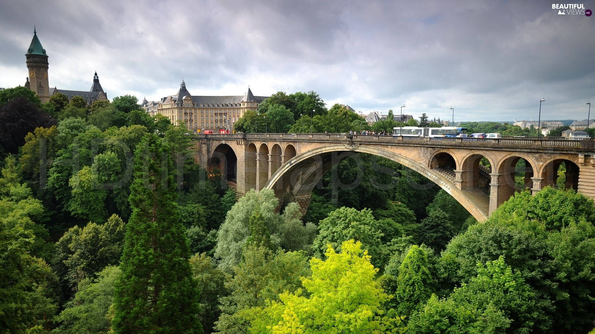 Luxembourg 桌布 壁紙 — 一座宏偉的石拱橋橫跨鬱鬱蔥蔥的綠色山谷，背後有歷史建築和城堡塔樓在陰天天空下聳立。