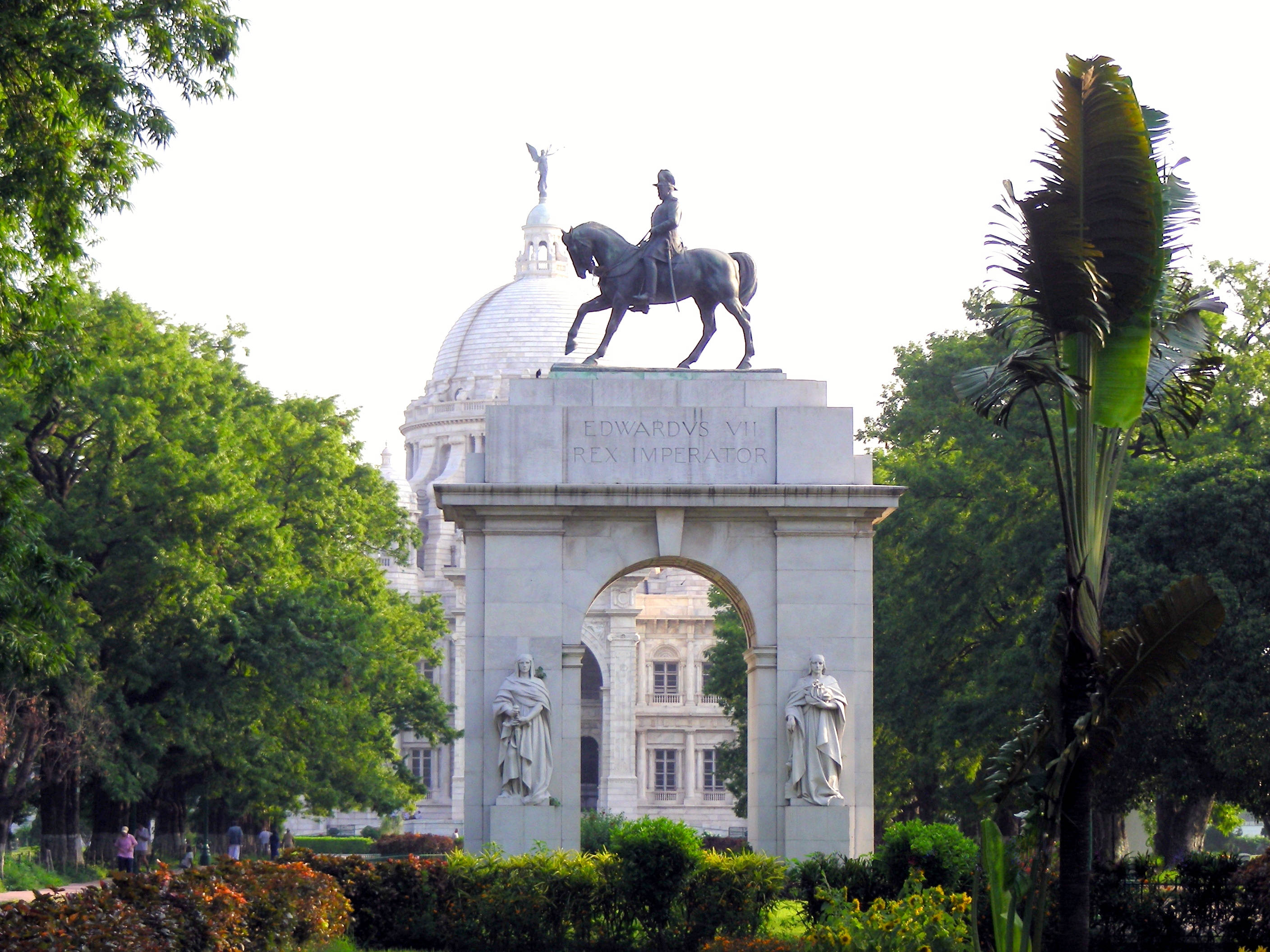 Kolkata 桌布 壁紙 — 一座宏偉的石拱門，頂部有愛德華七世的青銅騎馬雕像，矗立在茂密的綠樹和修剪整齊的樹籬中，沐浴在明亮的日光下。