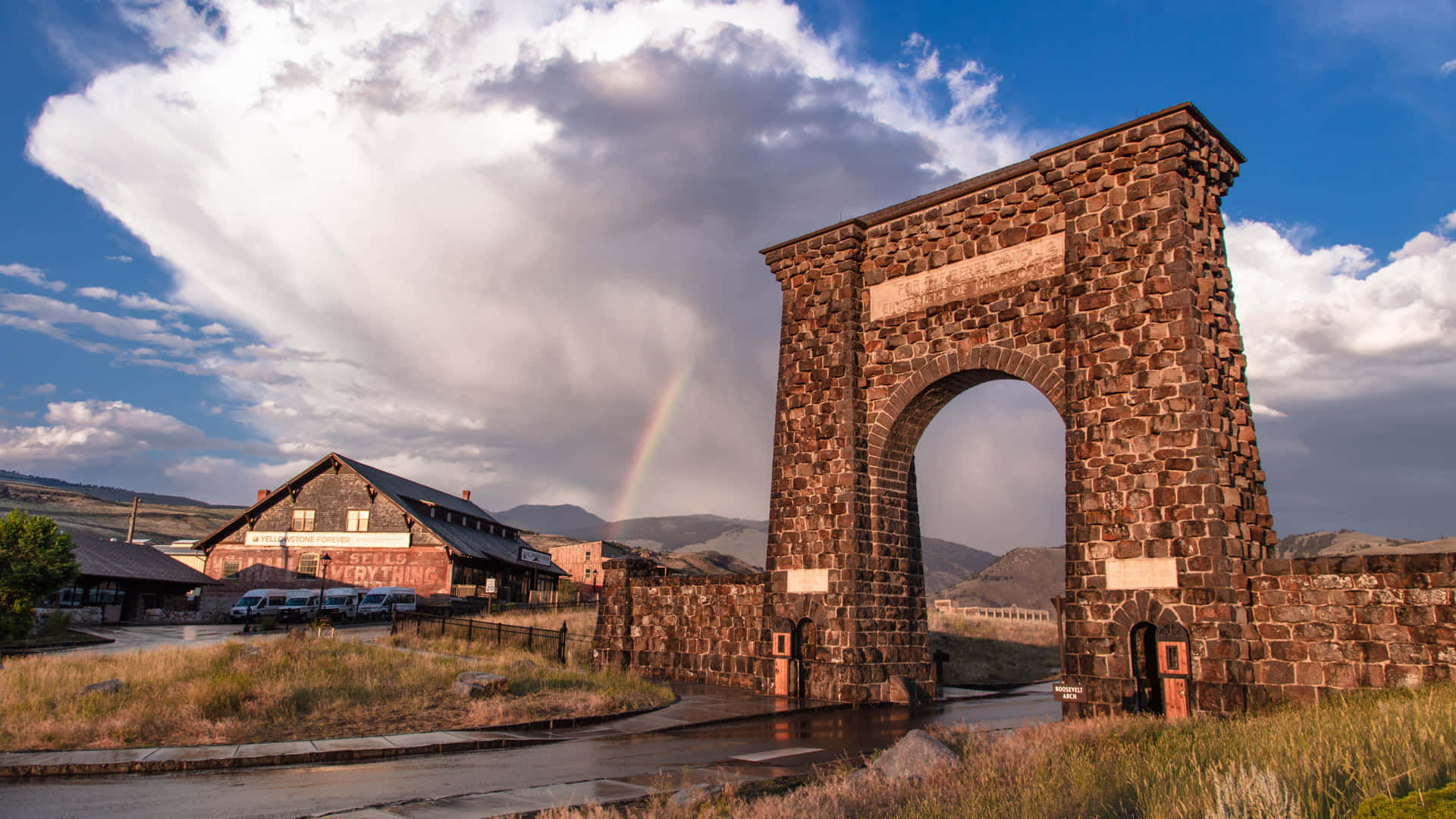 Yellowstone 桌布 壁紙 — 一座宏偉的石拱門在戲劇性的天空下高聳，背後有一道鮮豔的彩虹彎曲，附近有一座質樸的建築。