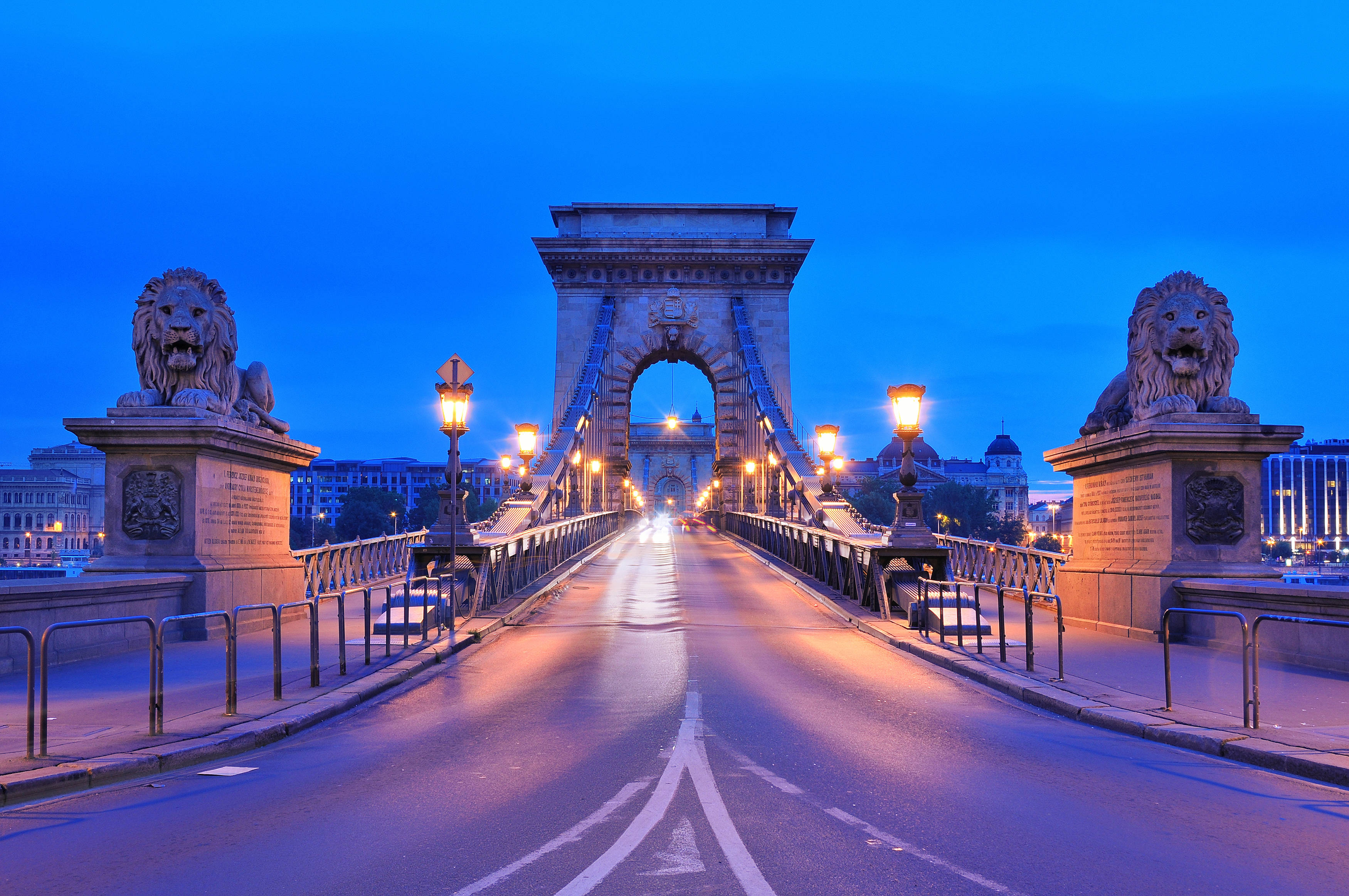 Hungary 桌布 壁紙 — 一座宏偉的石橋，上有獅子雕像和發光的燈，在深藍色的暮色天空下延伸於水面上。