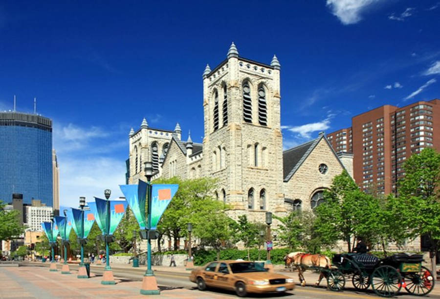 Minneapolis 桌布 壁紙 — 一座宏偉的石砌教堂，擁有高聳的塔樓，矗立在充滿活力的城市廣場上，藍天白雲下，附近有現代摩天大樓和馬車。