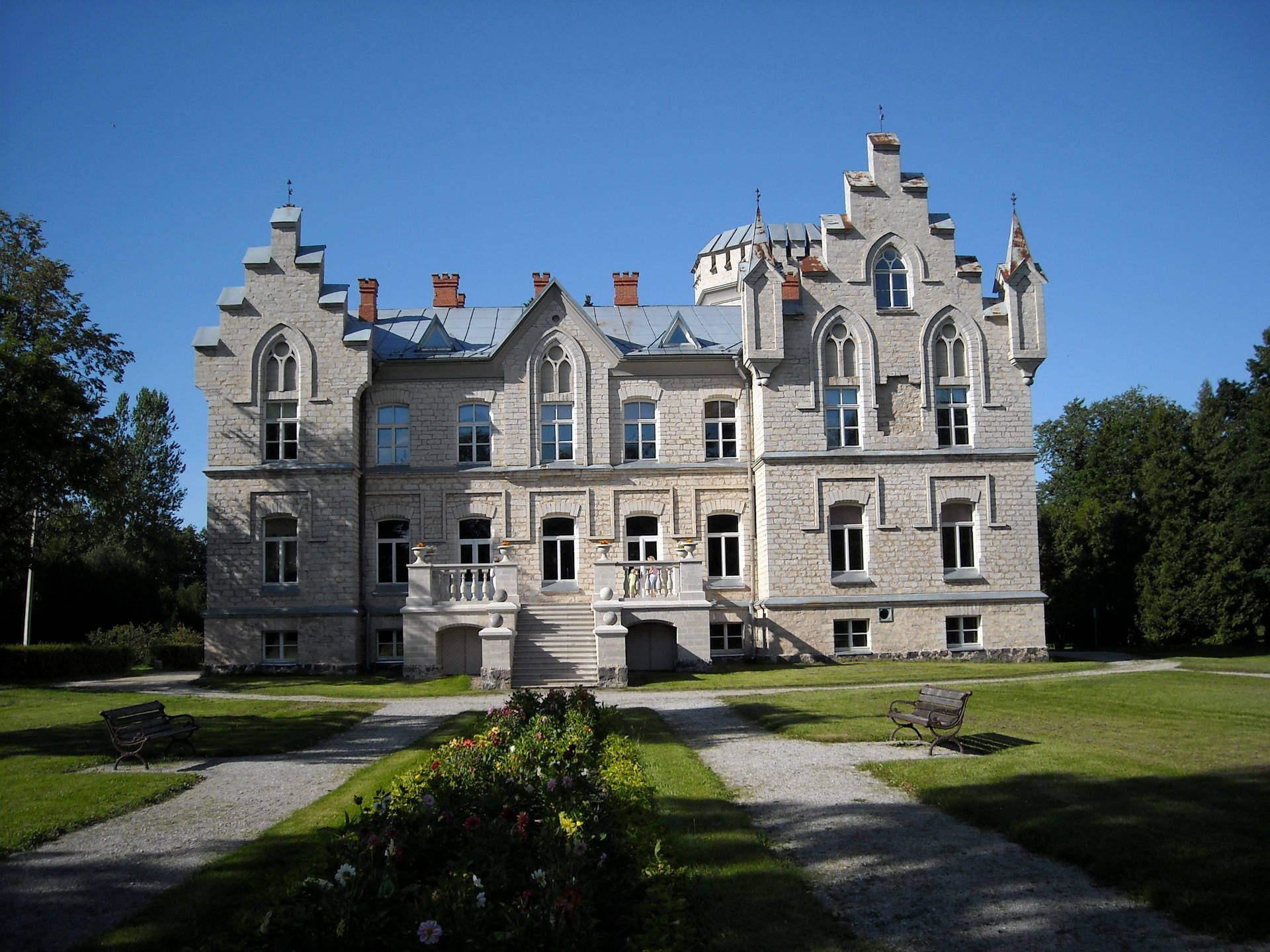Estonia 桌布 壁紙 — 一座宏偉的石砌莊園，帶有哥德式塔樓和拱形窗戶，矗立在晴朗的藍天下，周圍是修剪整齊的草坪和碎石小徑。