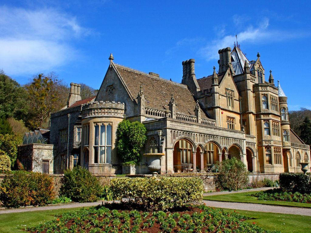 Victorian 桌布 壁紙 — 一座宏偉的石砌莊園，擁有塔樓和拱形窗戶，矗立在明亮的藍天下，周圍是修剪整齊的花園和秋色樹木。
