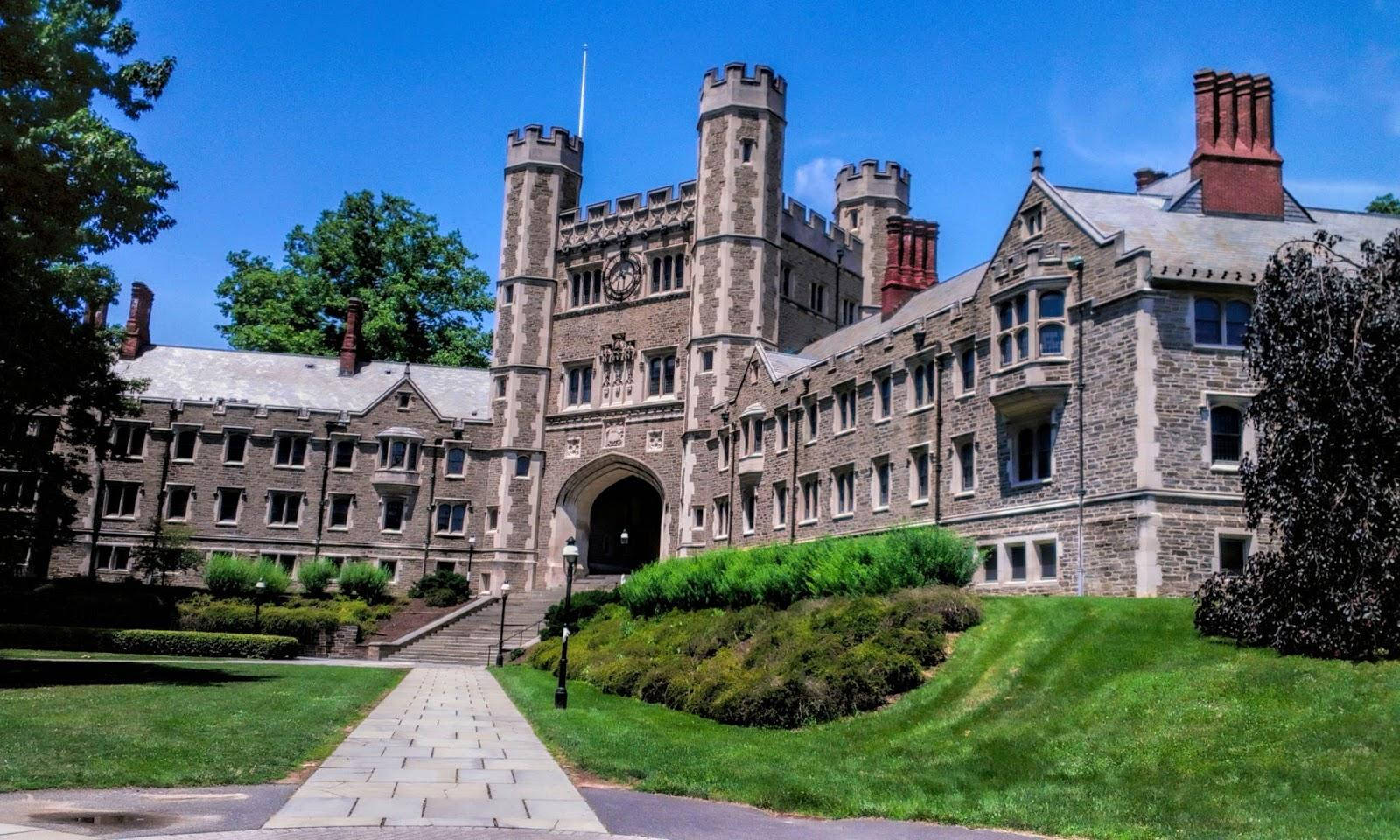 學校 桌布 壁紙 — 一座宏偉的石造城堡風格大學建築，設有塔樓和拱形入口，矗立在晴朗的藍天下，兩旁是茂密的綠色草坪和成熟的樹木。
