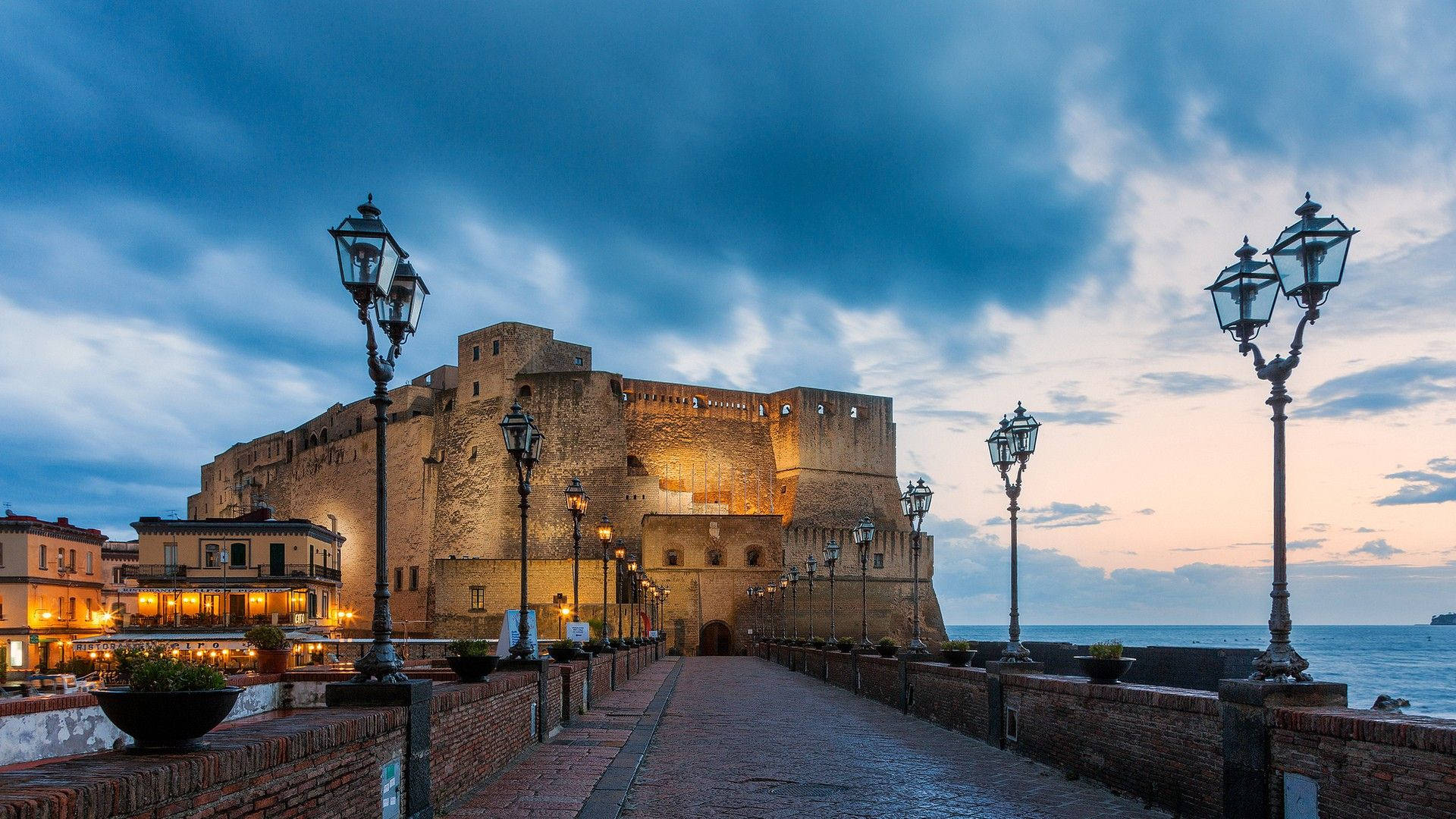 Naples 桌布 壁紙 — 一座宏偉的石造堡壘在黃昏時分被照亮，復古街燈排列在海邊的鵝卵石小徑旁。