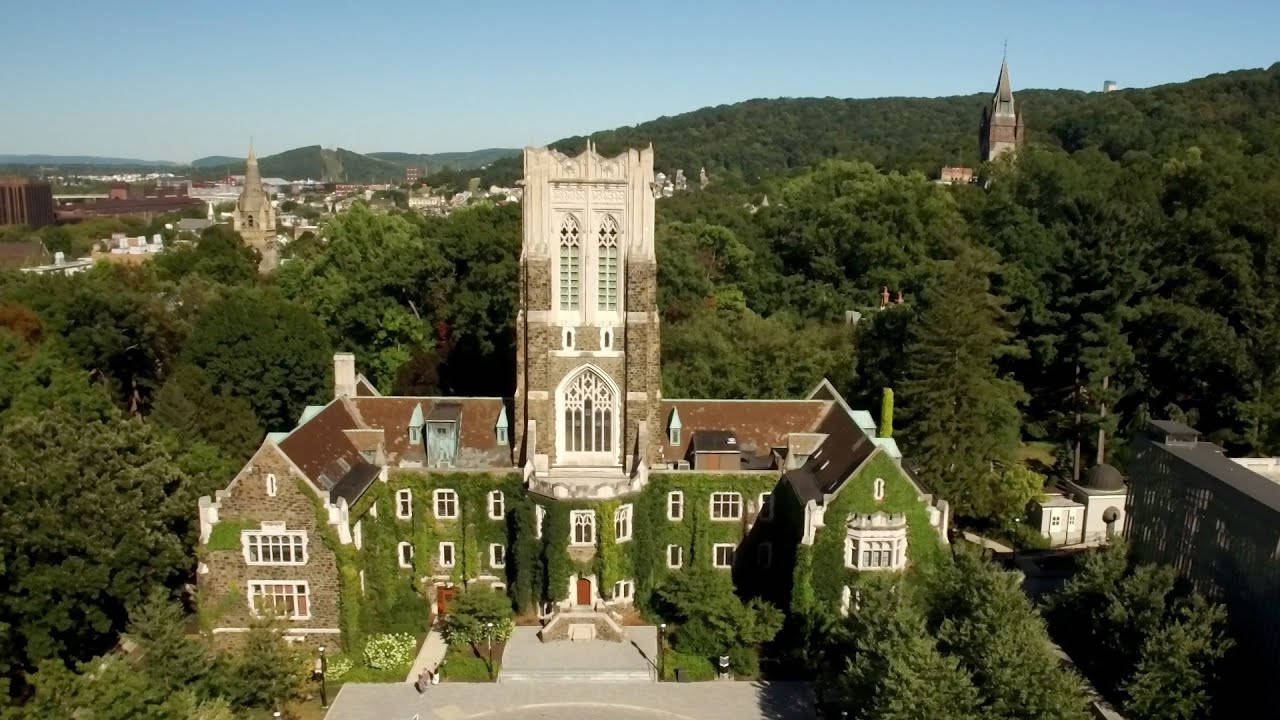 Building 桌布 壁紙 — 一座宏偉的石造大學建築的鳥瞰圖，牆壁爬滿常春藤，高聳的哥德式塔樓周圍環繞著茂密的綠樹，背景是清澈的藍天。