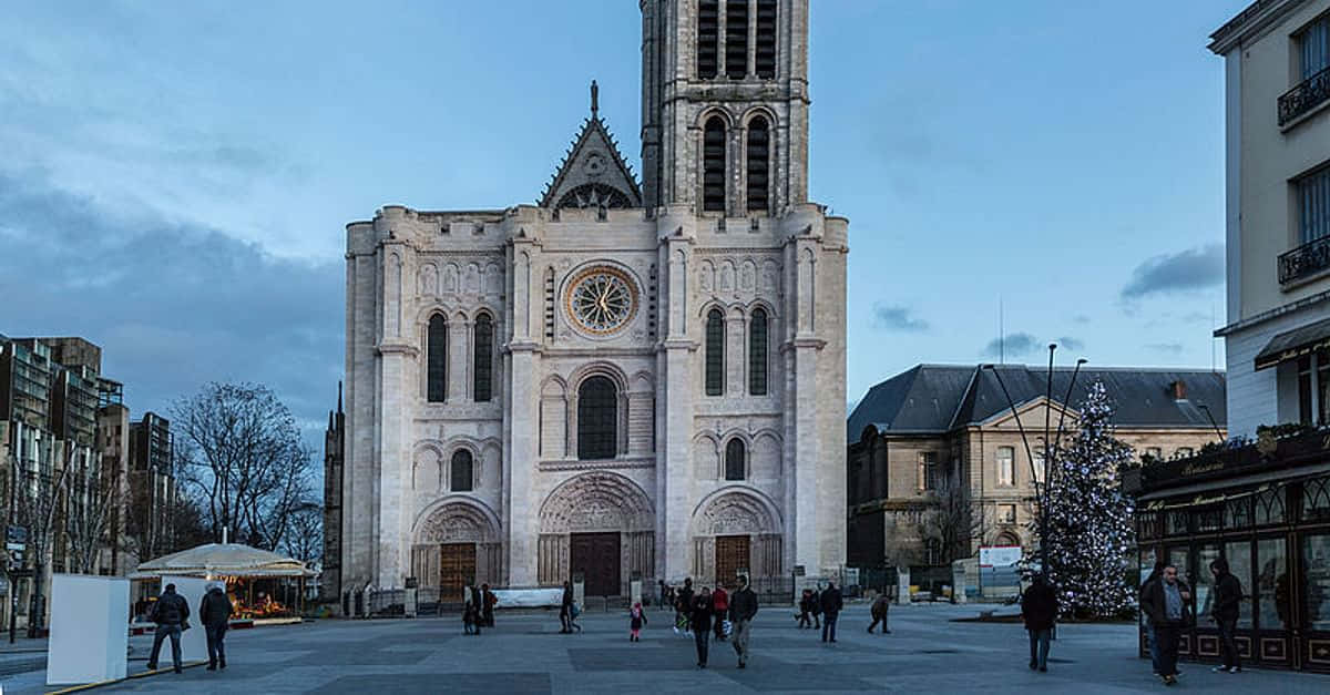 Gothic Architecture 桌布 壁紙 — 一座宏偉的石造大教堂，帶有玫瑰窗，矗立在黃昏的城市廣場上，人們漫步經過裝飾好的聖誕樹。