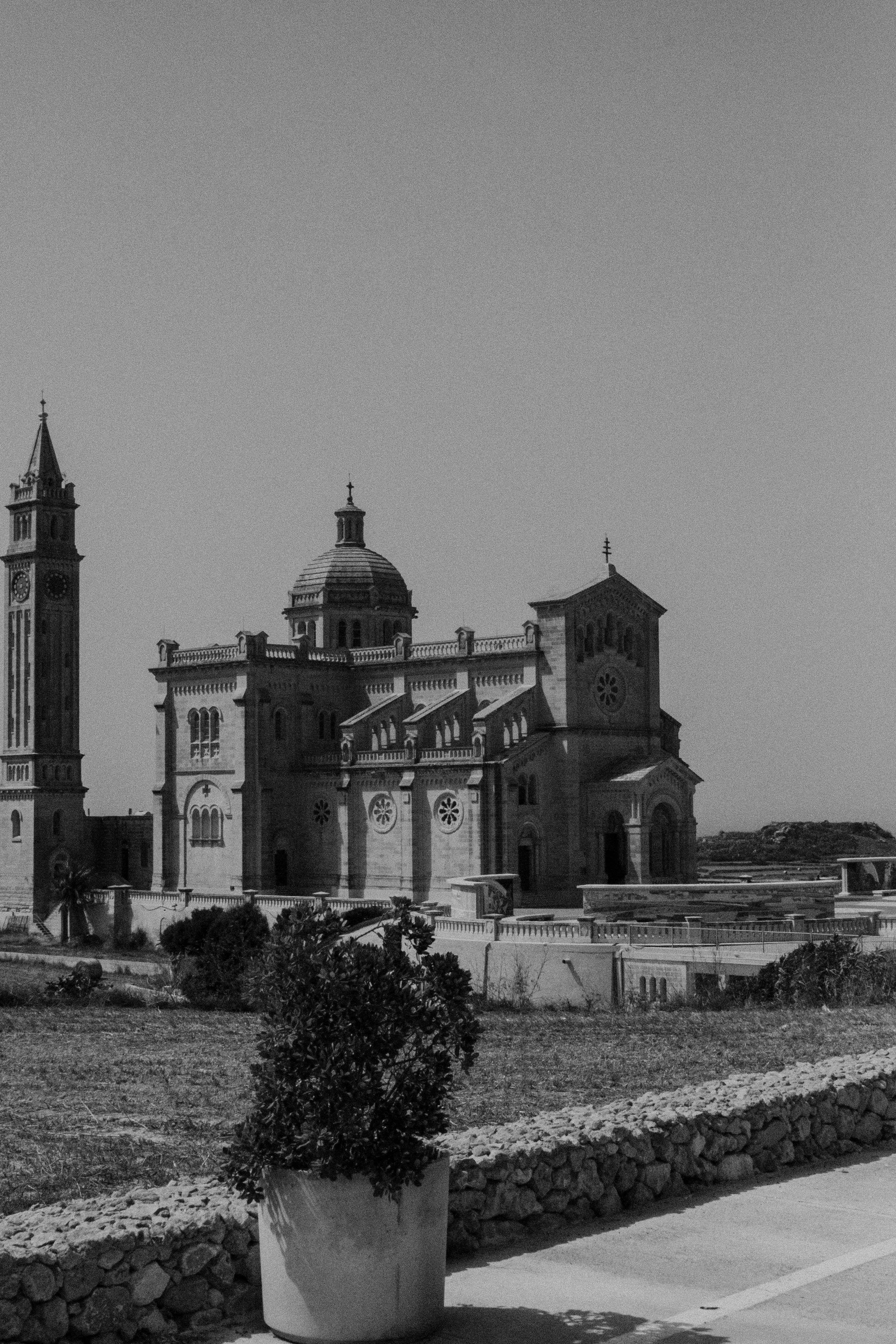 Malta 桌布 壁紙 — 一座宏偉的石造大教堂，帶有鐘樓和圓頂，在晴朗的天空下莊嚴地矗立，由盆栽植物和石牆以單色構圖框住。