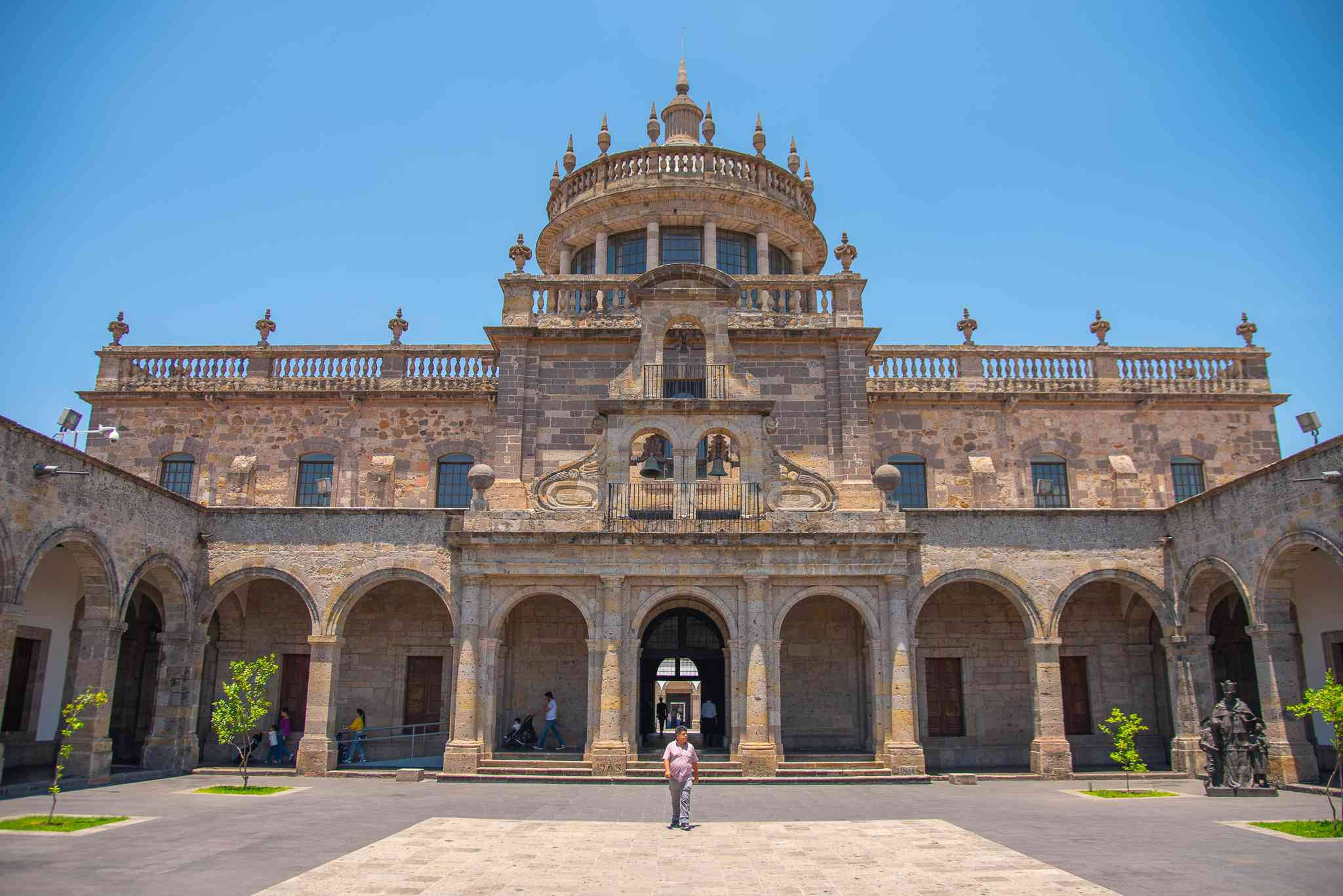 Guadalajara 桌布 壁紙 — 一座宏偉的石造大教堂，擁有拱形柱廊和圓頂塔樓，矗立在晴朗的藍天下，庭院裡有幾位訪客。