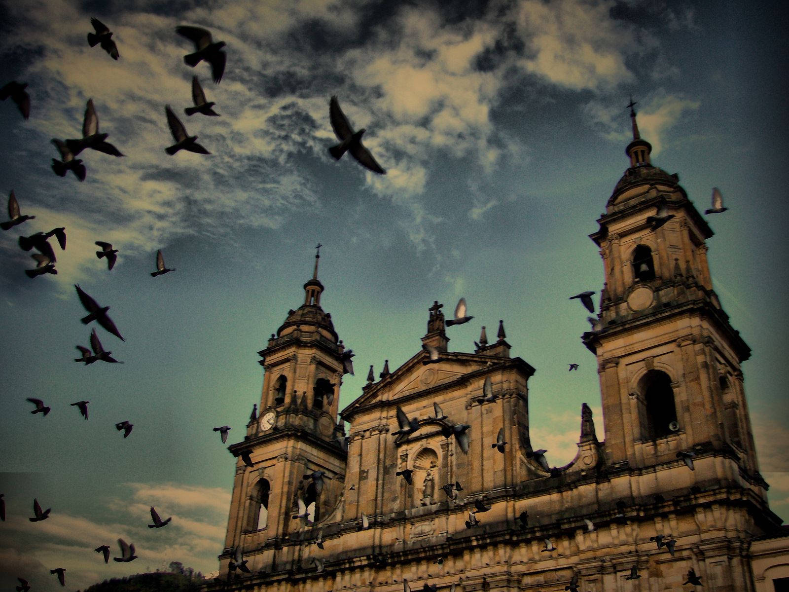 Bogota 桌布 壁紙 — 一座宏偉的石造大教堂，擁有雙塔，矗立在戲劇性的多雲天空下，鳥兒在其古老的外牆周圍飛翔和棲息。