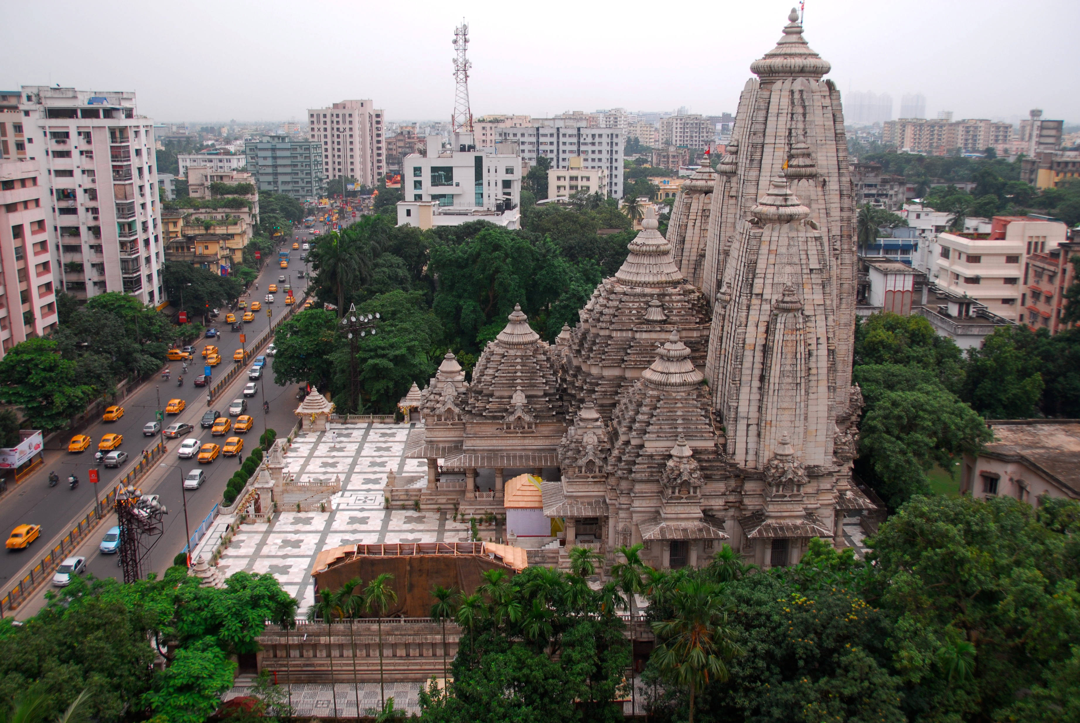 Kolkata 桌布 壁紙 — 一座宏偉的石造寺廟，擁有精緻的尖塔，聳立在充滿黃色計程車和現代建築的繁華城市街道之上。