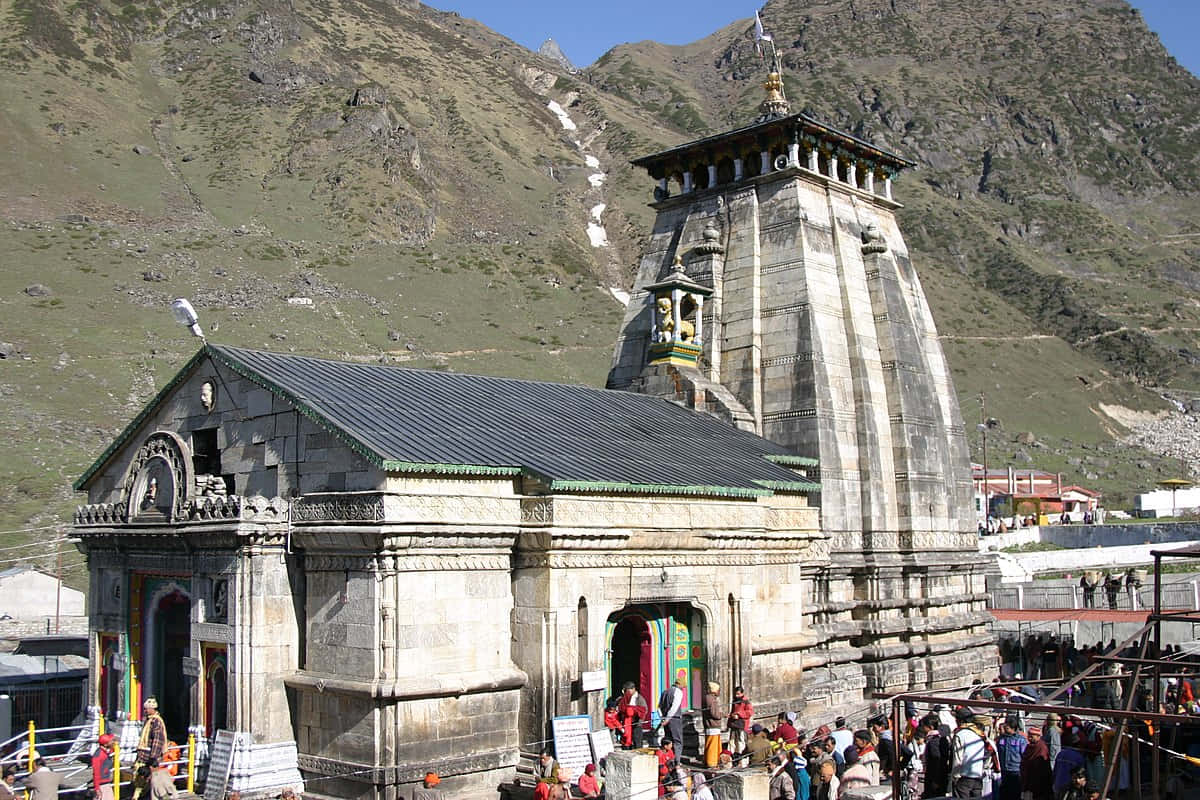 Kedarnath 桌布 壁紙 — 一座宏偉的石造寺廟，擁有高聳的尖塔，坐落在崎嶇的山脈中，在清澈的藍天下，入口處聚集了朝聖者。