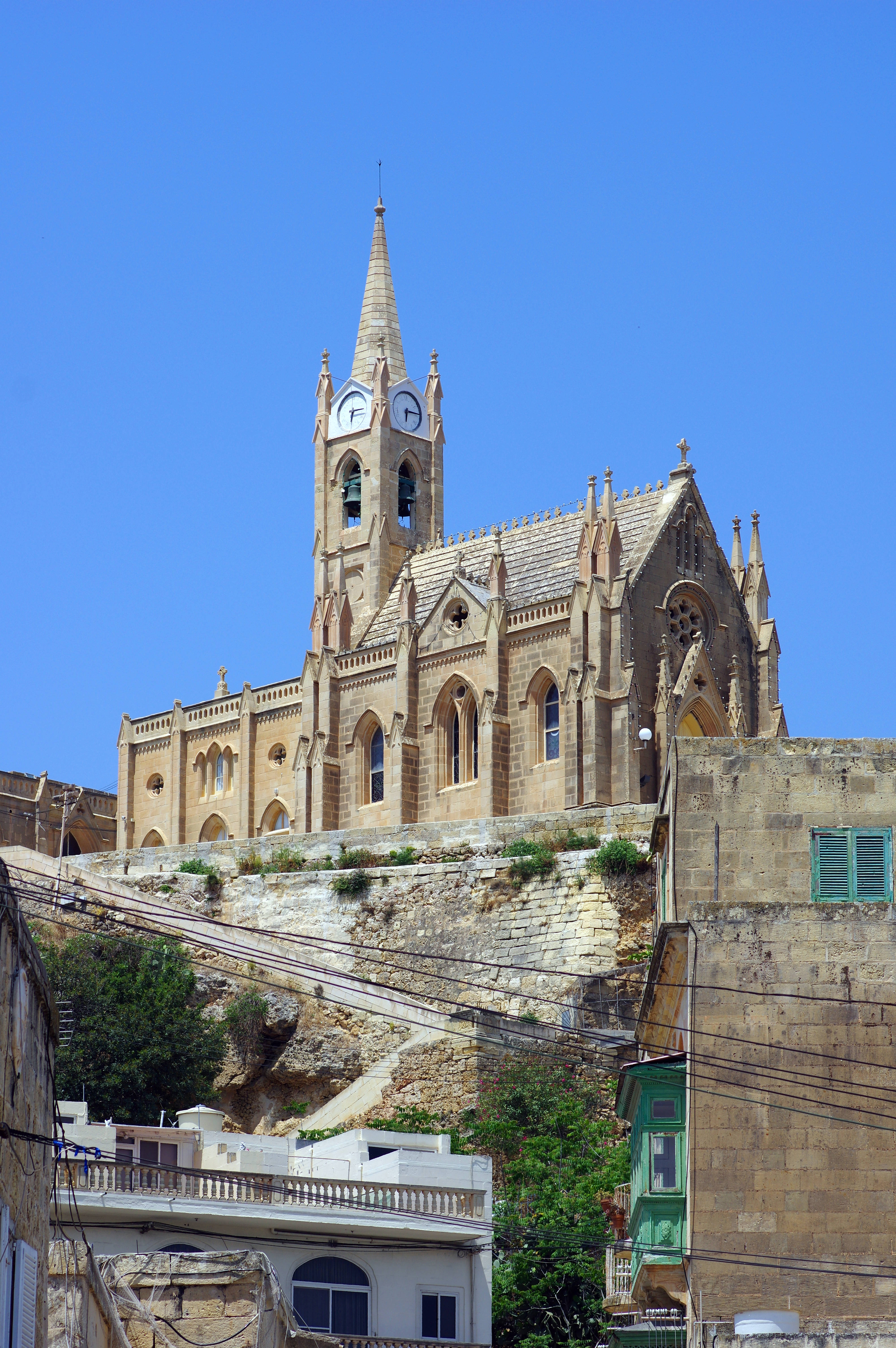 Malta 桌布 壁紙 — 一座宏偉的石造教堂，高聳的尖塔在晴朗的藍天下，矗立於陽光普照的建築物之上。