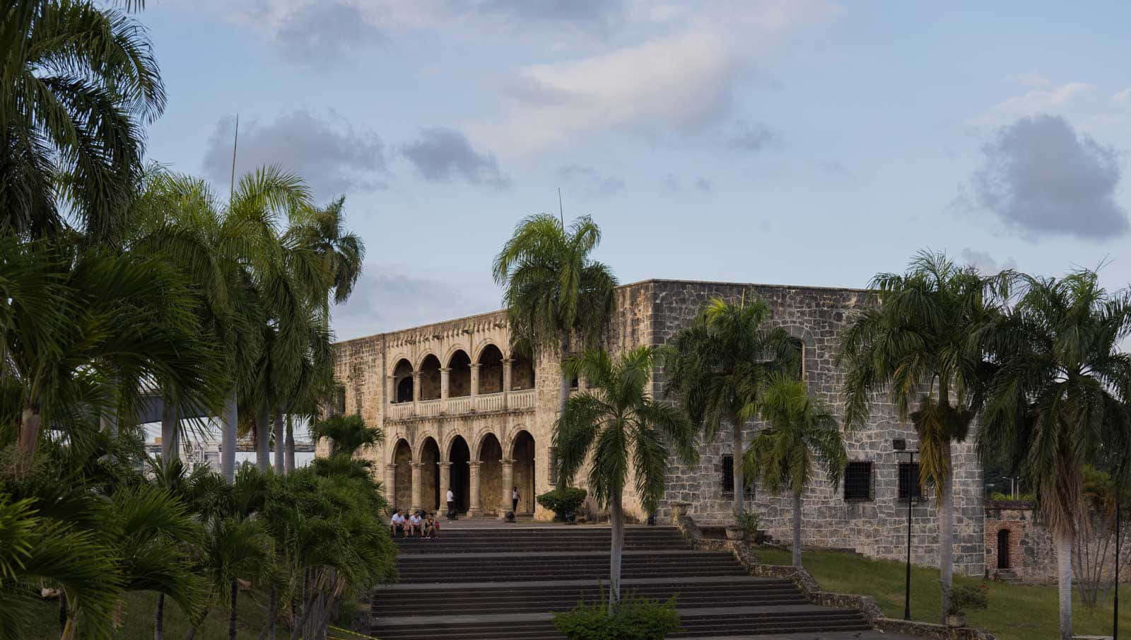 Museum 桌布 壁紙 — 一座宏偉的石造殖民風格建築，擁有拱形步道和棕櫚樹，矗立在多雲的天空下，階梯上有訪客。