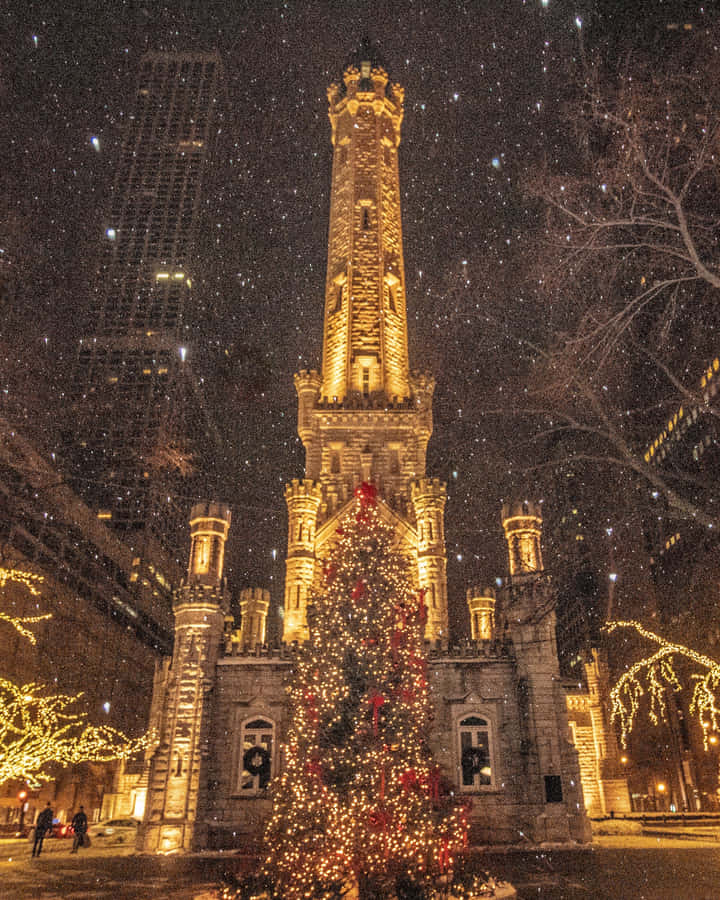 Aesthetic Christmas Tree 桌布 壁紙 — 一座宏偉的石造消防局塔樓在飄雪下散發溫暖光芒，周圍環繞著掛滿燈飾和紅色蝴蝶結的節日聖誕樹。
