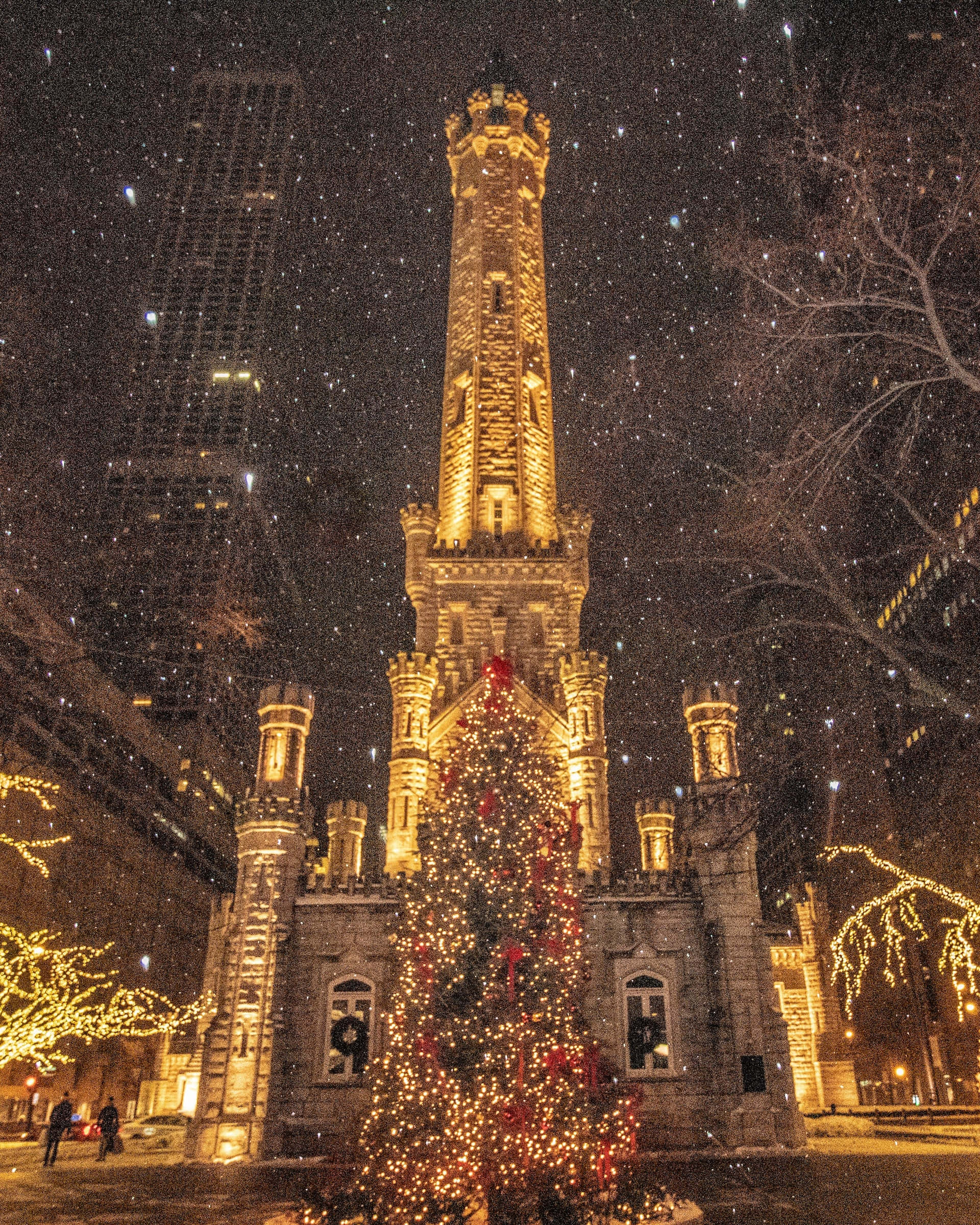 Aesthetic Christmas Tree 桌布 壁紙 — 一座宏偉的石造消防局塔樓在飄雪中溫暖地發光，周圍環繞著掛滿燈飾和紅色蝴蝶結的節日聖誕樹。