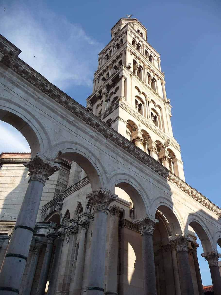 Croatia 桌布 壁紙 — 一座宏偉的石造鐘樓聳立於柱廊拱門之上，在清澈的藍天下，柔和陽光投下長長的影子。