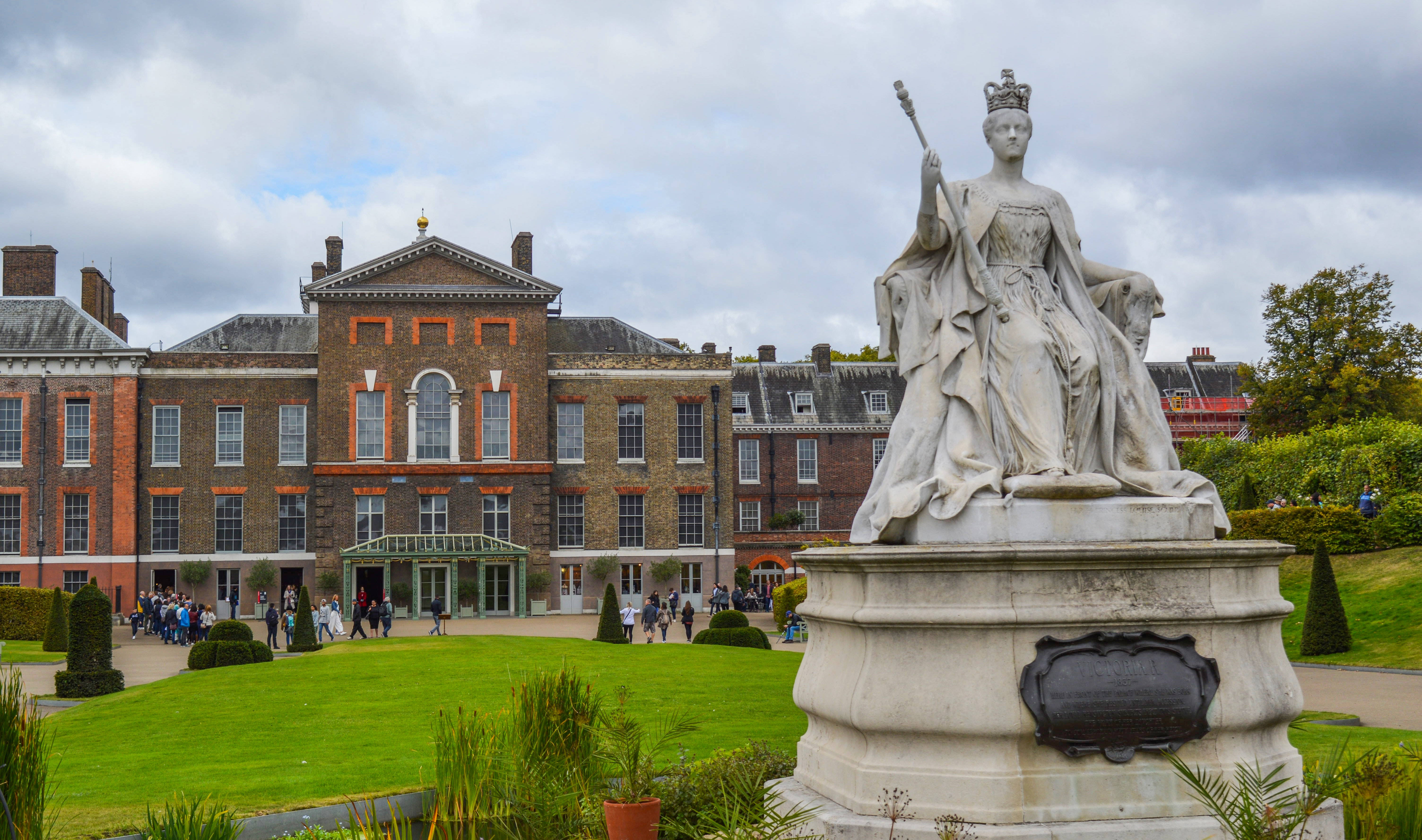 England 桌布 壁紙 — 一座宏偉的石雕女王雕像，頭戴王冠，手持權杖，矗立在修剪整齊的花園中，背後是歷史悠久的磚造宮殿，天空多雲。