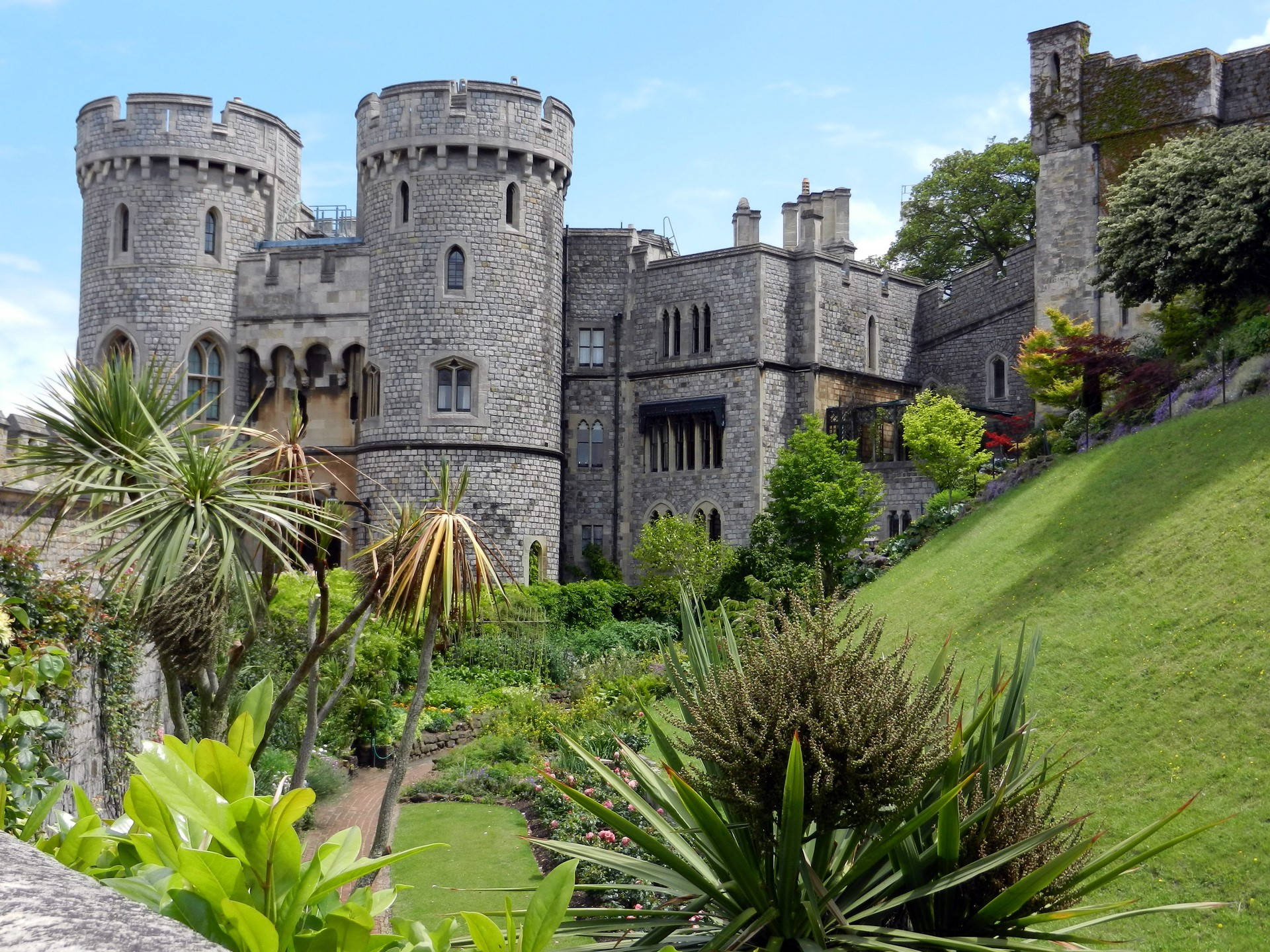 城堡動畫 桌布 壁紙 — 一座宏偉的石頭城堡，帶有塔樓和拱形窗戶，聳立在鬱鬱蔥蔥的綠色花園和熱帶植物之上，天空清澈湛藍。