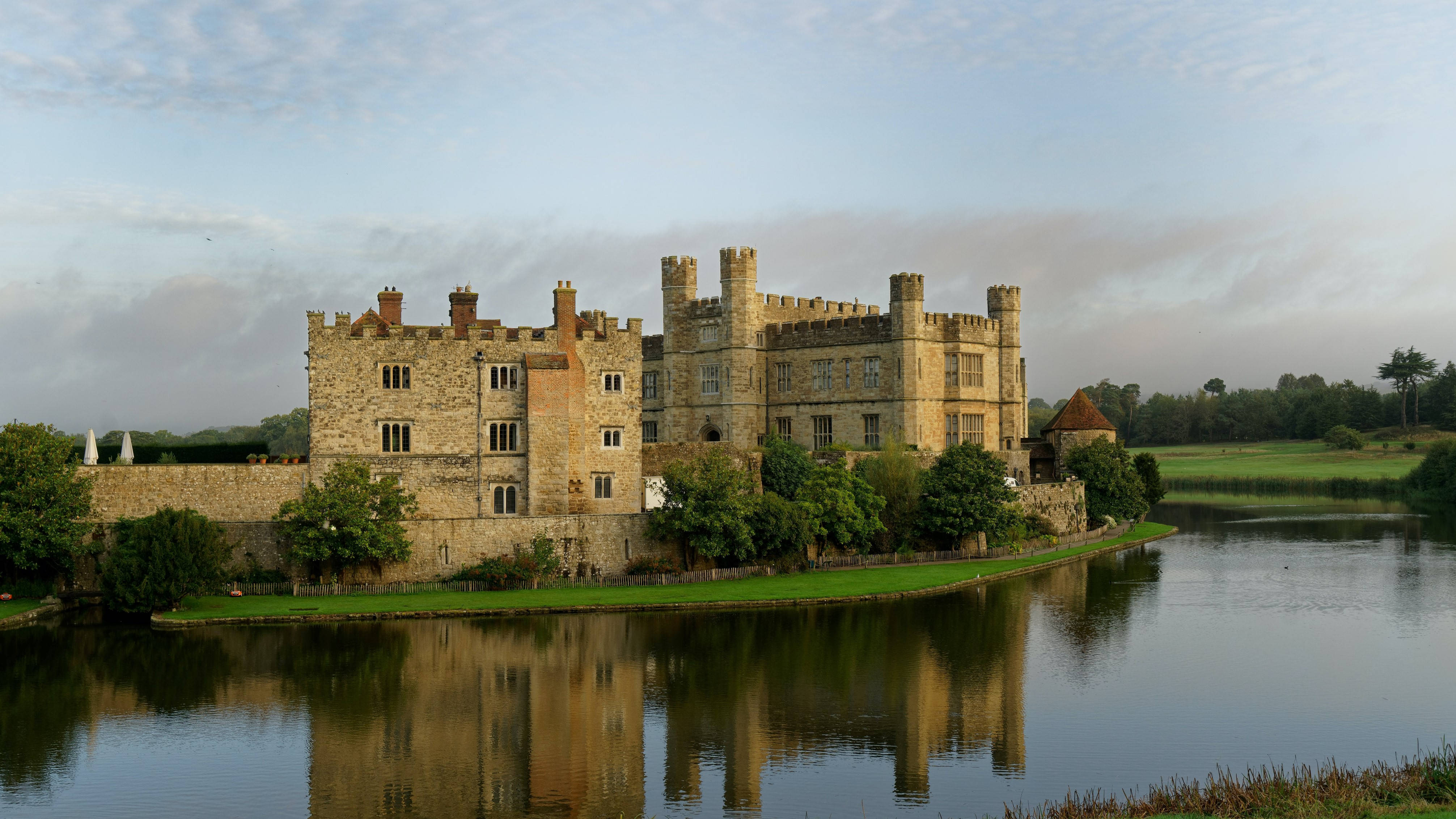 England 桌布 壁紙 — 一座宏偉的石頭城堡，帶有塔樓和煙囪，矗立在平靜的湖邊，在柔和的多雲天空下，湖面倒映著它的輪廓。
