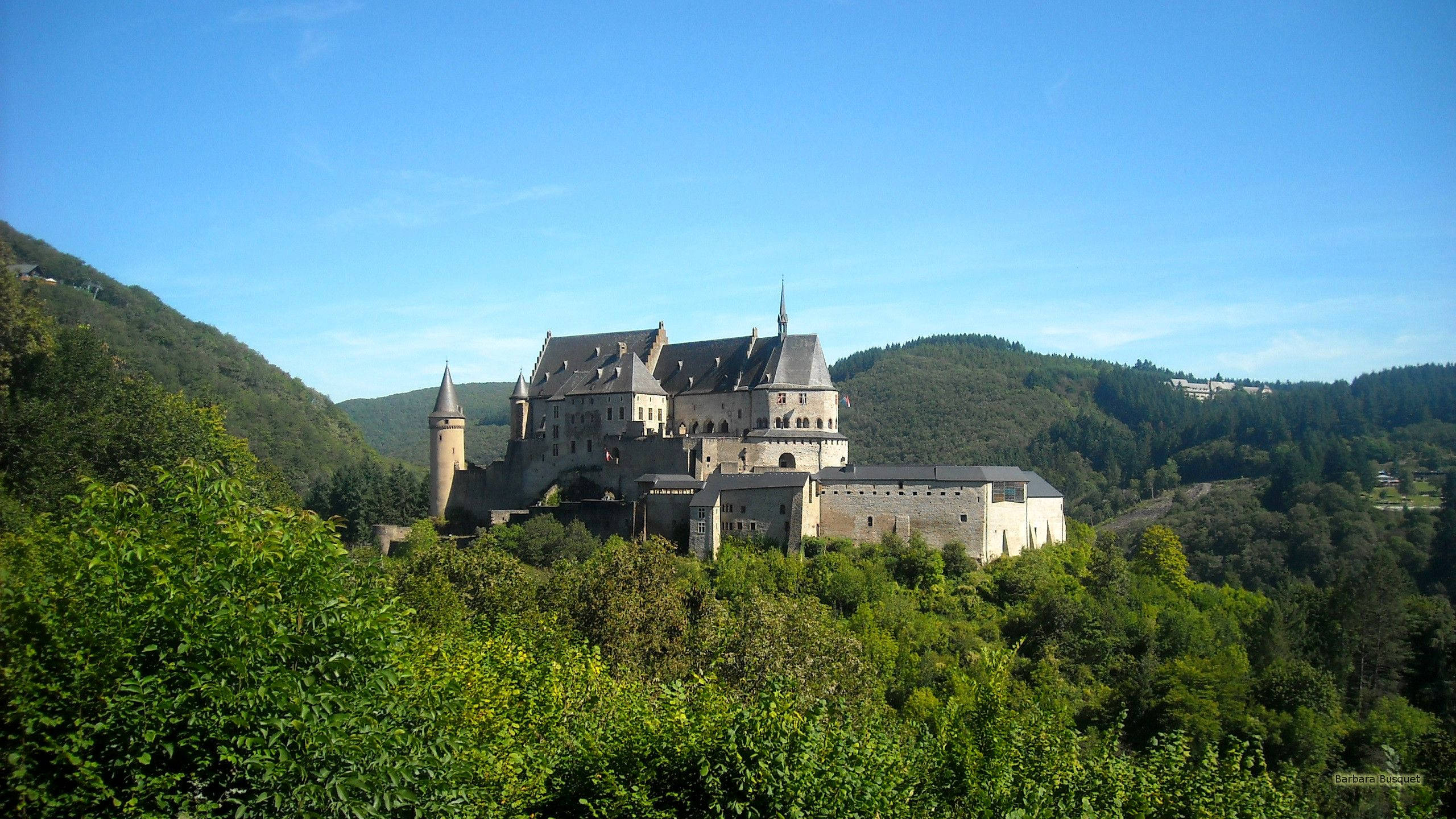 Luxembourg 桌布 壁紙 — 一座宏偉的石頭城堡，擁有塔樓和尖頂，坐落在鬱鬱蔥蔥的綠色山丘上，在清澈的藍天下。