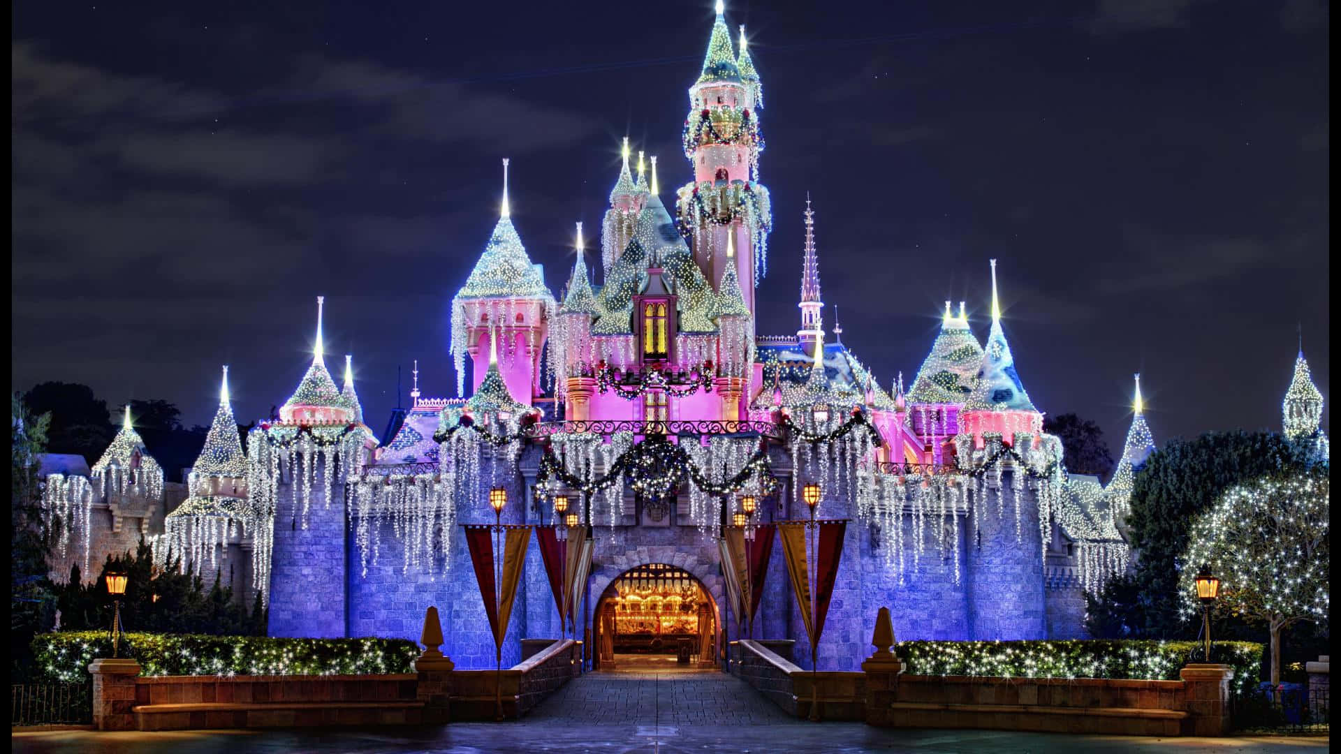 1920x1080 Disney 桌布 壁紙 — 一座宏偉的童話城堡，在漆黑的夜空下被鮮豔的粉紅色、藍色和白色燈光點亮。