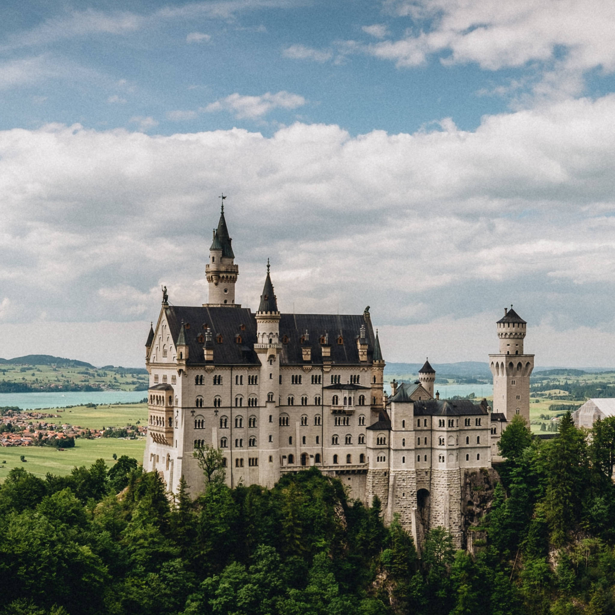 Neuschwanstein Castle 桌布 壁紙 — 一座宏偉的童話城堡，帶有塔樓和深色屋頂，坐落在森林覆蓋的山丘上，俯瞰山谷和湖泊，天空部分多雲。