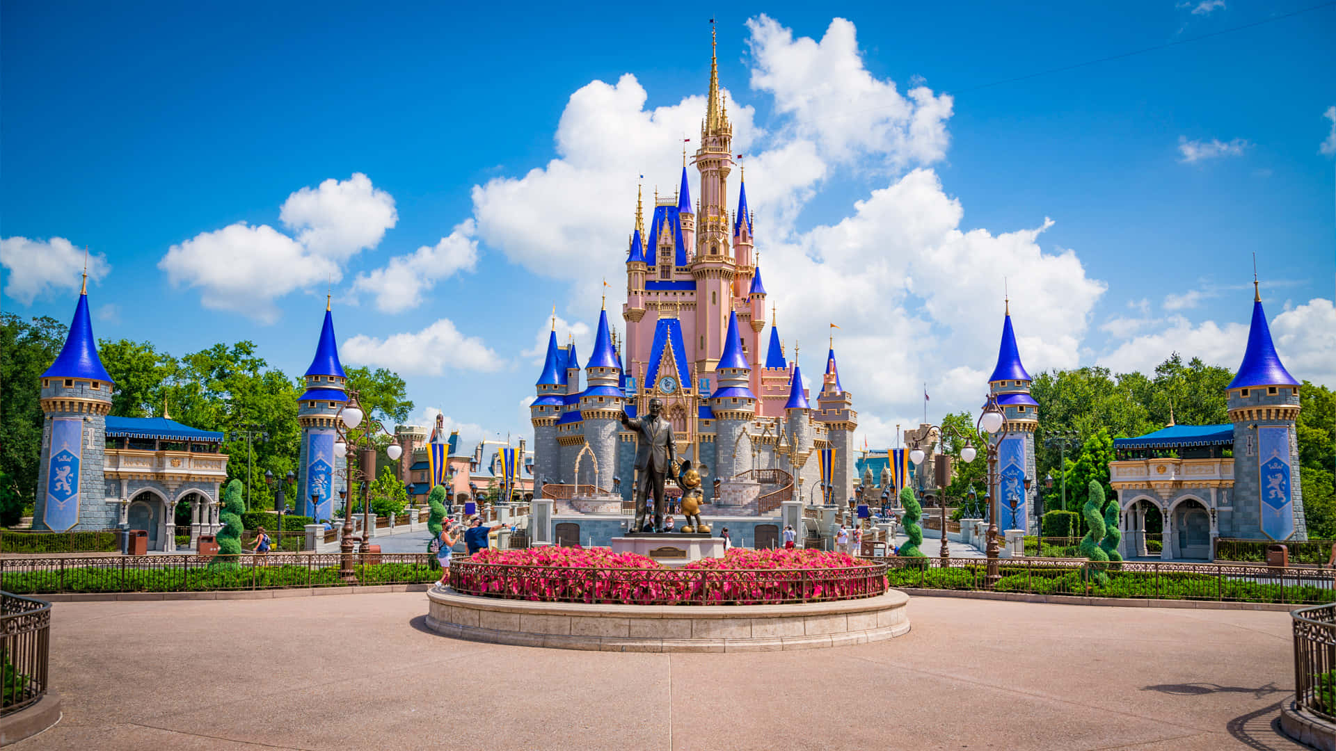 1920x1080 Disney 桌布 壁紙 — 一座宏偉的童話城堡，擁有藍色尖頂和金色點綴，矗立在晴朗的藍天下，飄著蓬鬆的白雲。