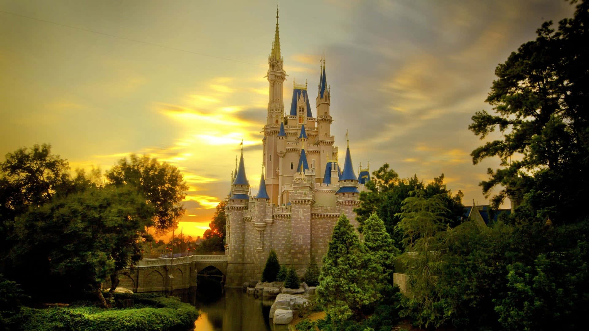 1920x1080 Disney 桌布 壁紙 — 一座宏偉的童話城堡，擁有藍色尖頂，矗立在日落時分，周圍環繞著茂密的綠樹和寧靜的護城河。