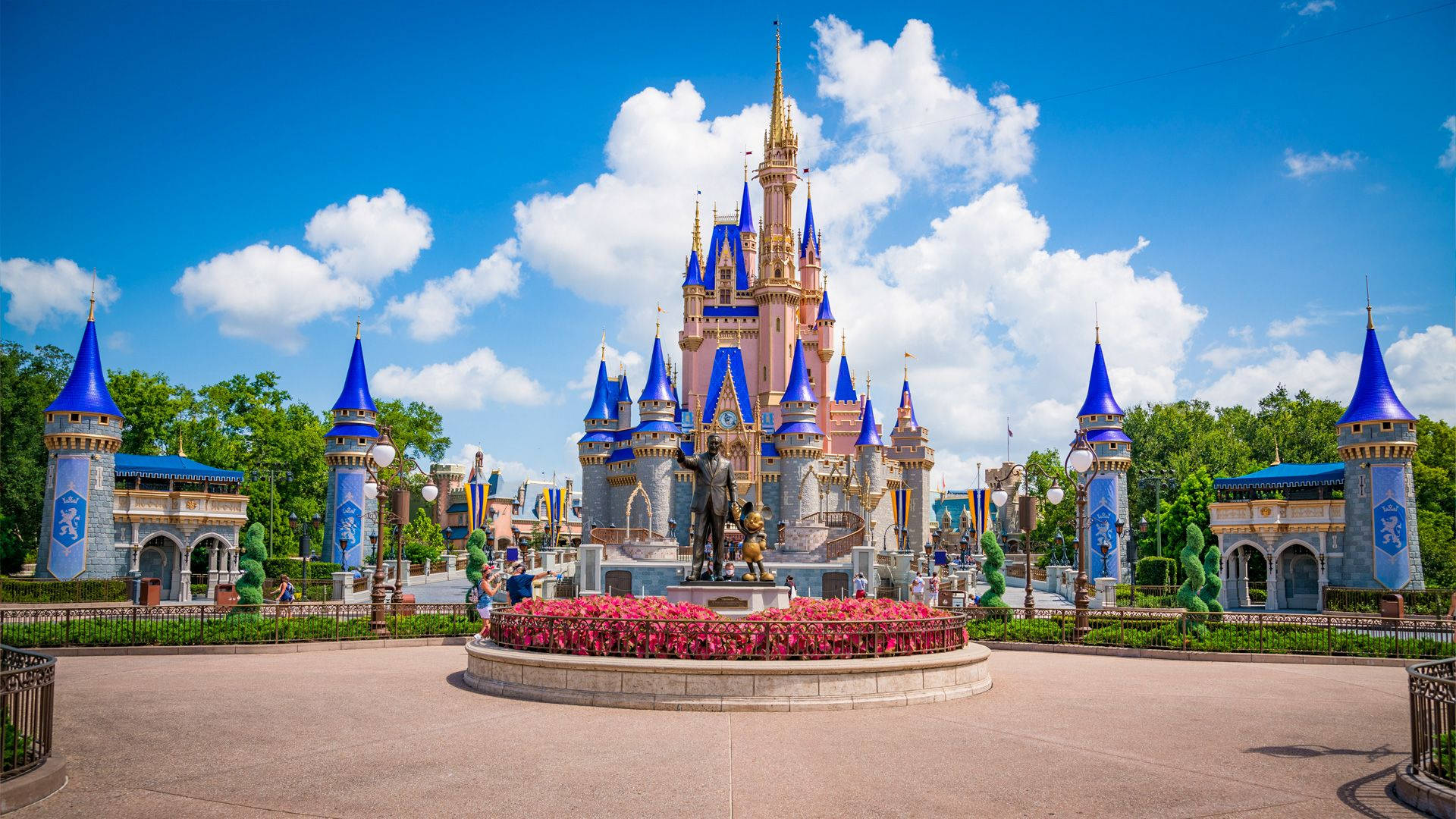 Disney Castle 桌布 壁紙 — 一座宏偉的童話城堡，擁有藍色尖頂和粉紅色石牆，矗立在點綴著蓬鬆白雲的明亮藍天下。