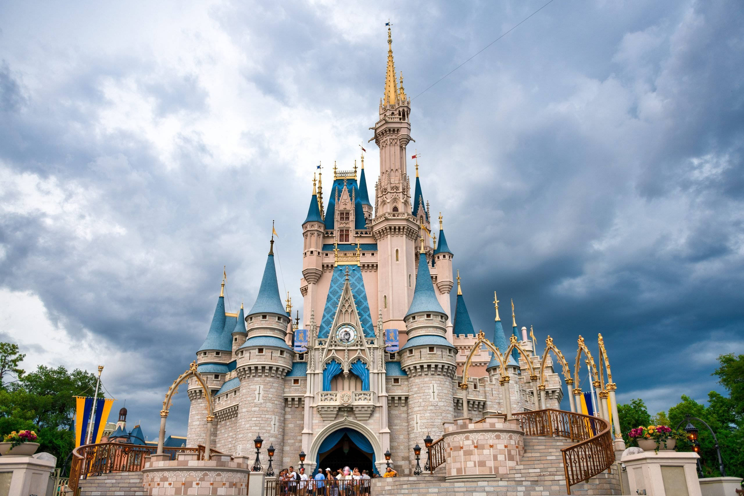 Disney Castle 桌布 壁紙 — 一座宏偉的童話城堡，擁有藍色尖頂和金色點綴，在戲劇性的多雲天空下巍然聳立。
