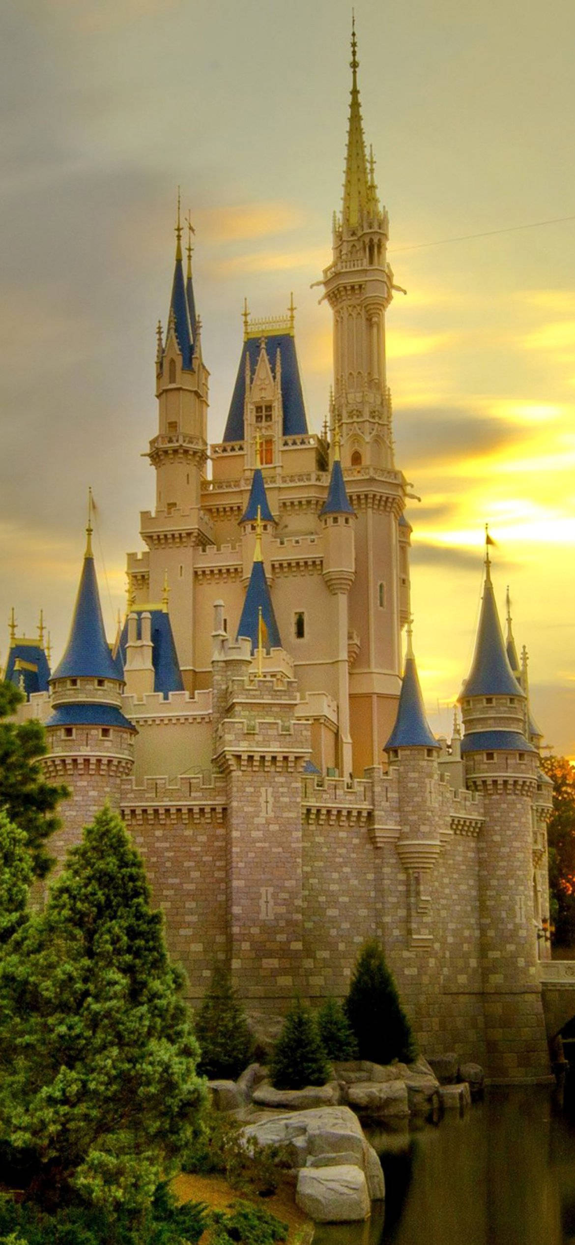 Disney Castle 桌布 壁紙 — 一座宏偉的童話城堡，擁有藍色尖頂和金色點綴，在夕陽天空下溫暖地閃耀。