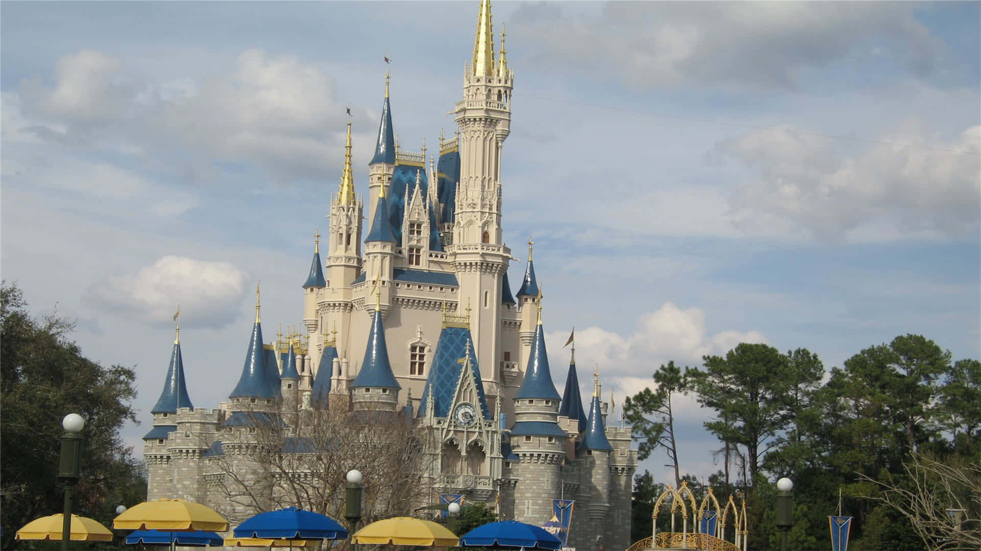 1920x1080 Disney 桌布 壁紙 — 一座宏偉的童話城堡，擁有藍色屋頂和金色尖塔，聳立在部分多雲的天空下，周圍環繞著樹木和彩色雨傘。
