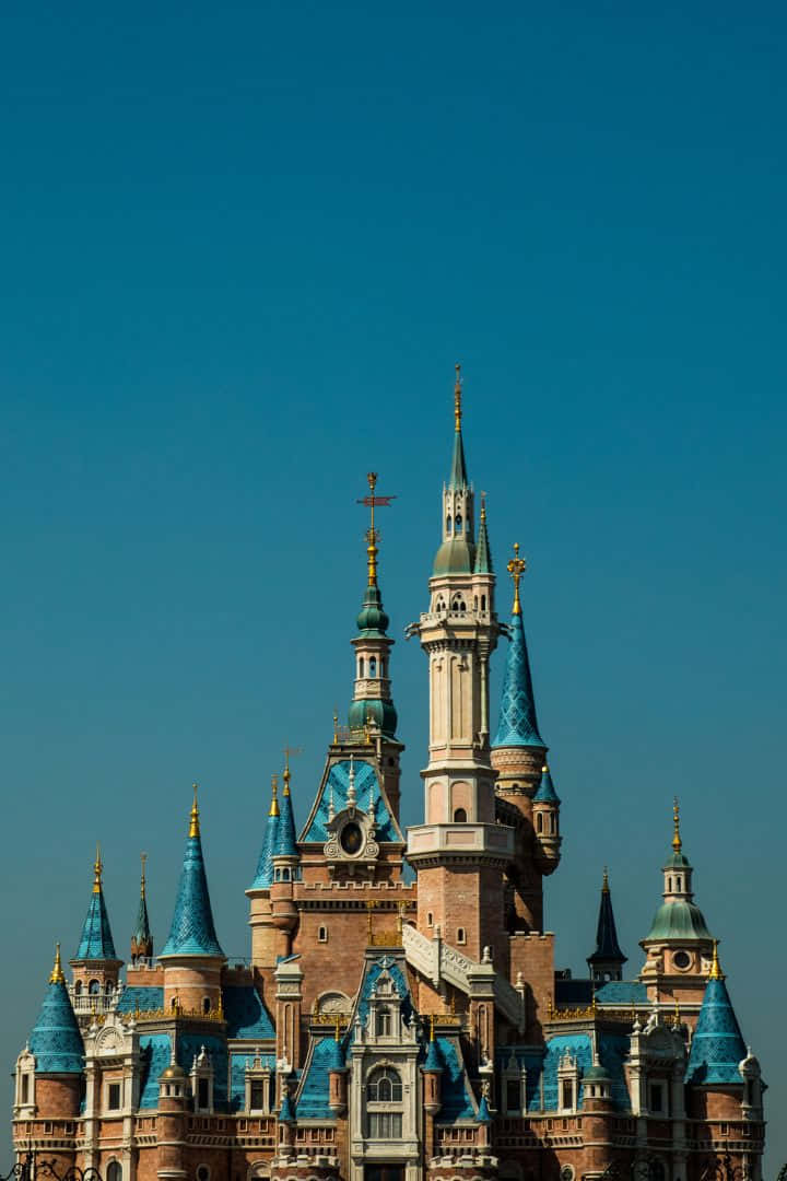 disneyland iPhone 桌布 壁紙 — 一座宏偉的童話城堡，擁有藍色瓷磚尖頂和金色點綴，在清澈的深藍色天空下聳立。