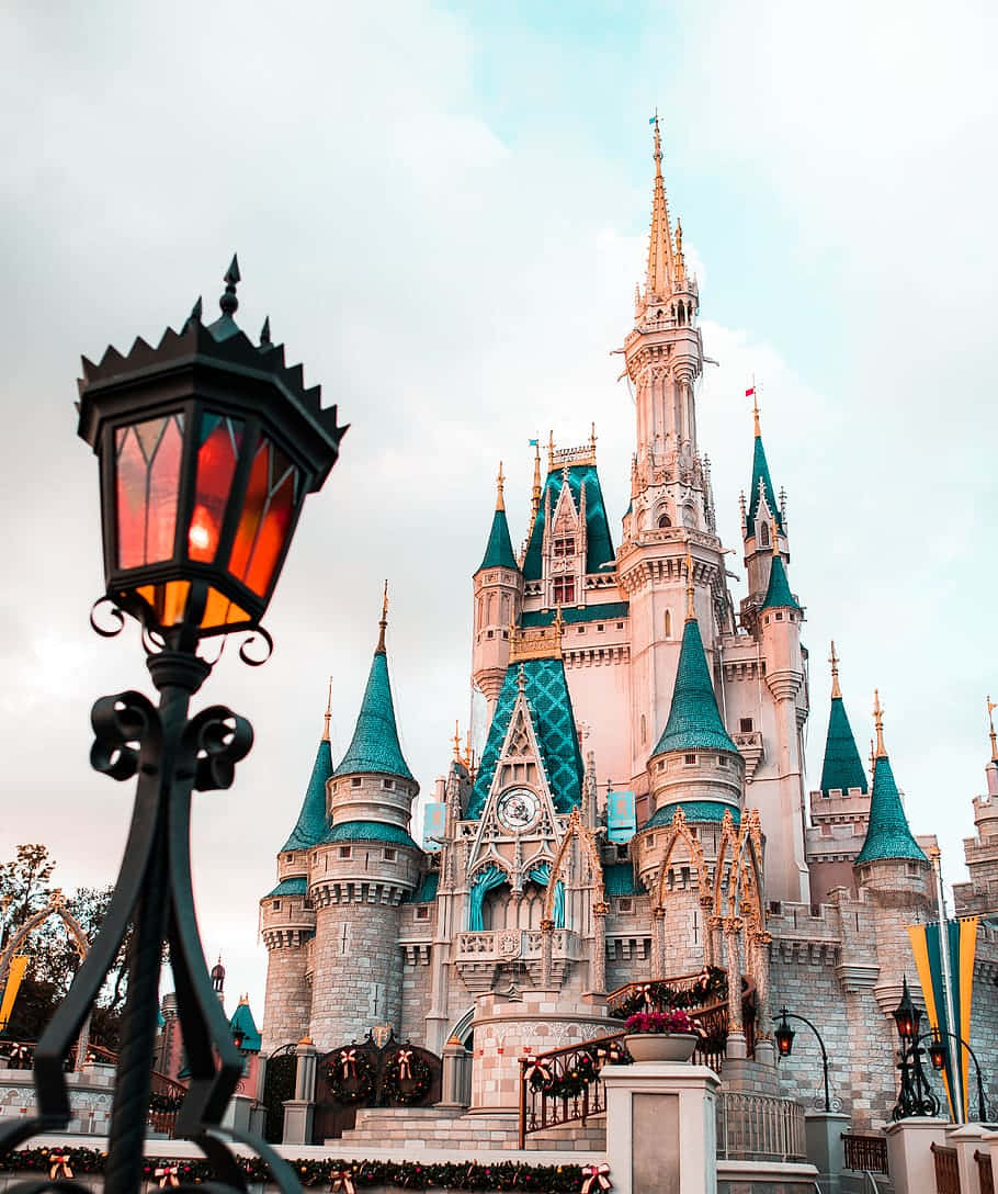Disney Castle 桌布 壁紙 — 一座宏偉的童話城堡，擁有青綠色的尖頂和金色點綴，在柔和天空下巍然聳立，前景中閃爍的復古燈籠將其框住。