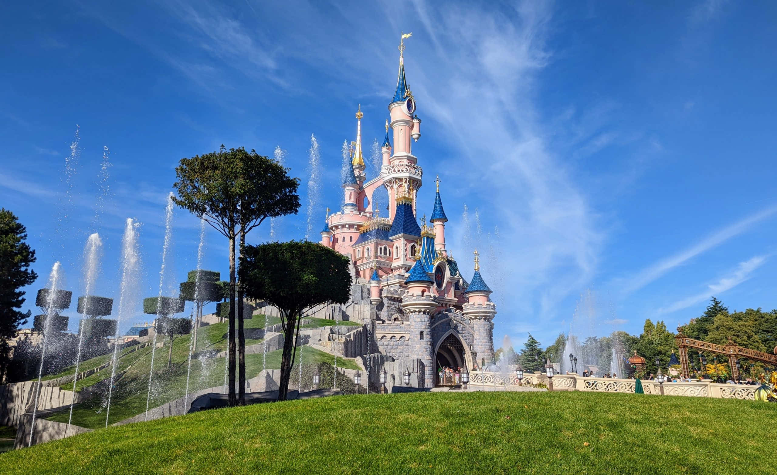 Disneyland Castle 桌布 壁紙 — 一座宏偉的粉紅色和藍色城堡，帶有尖塔，聳立在修剪整齊的草坪和活躍的噴泉之上，在明亮的藍天下。