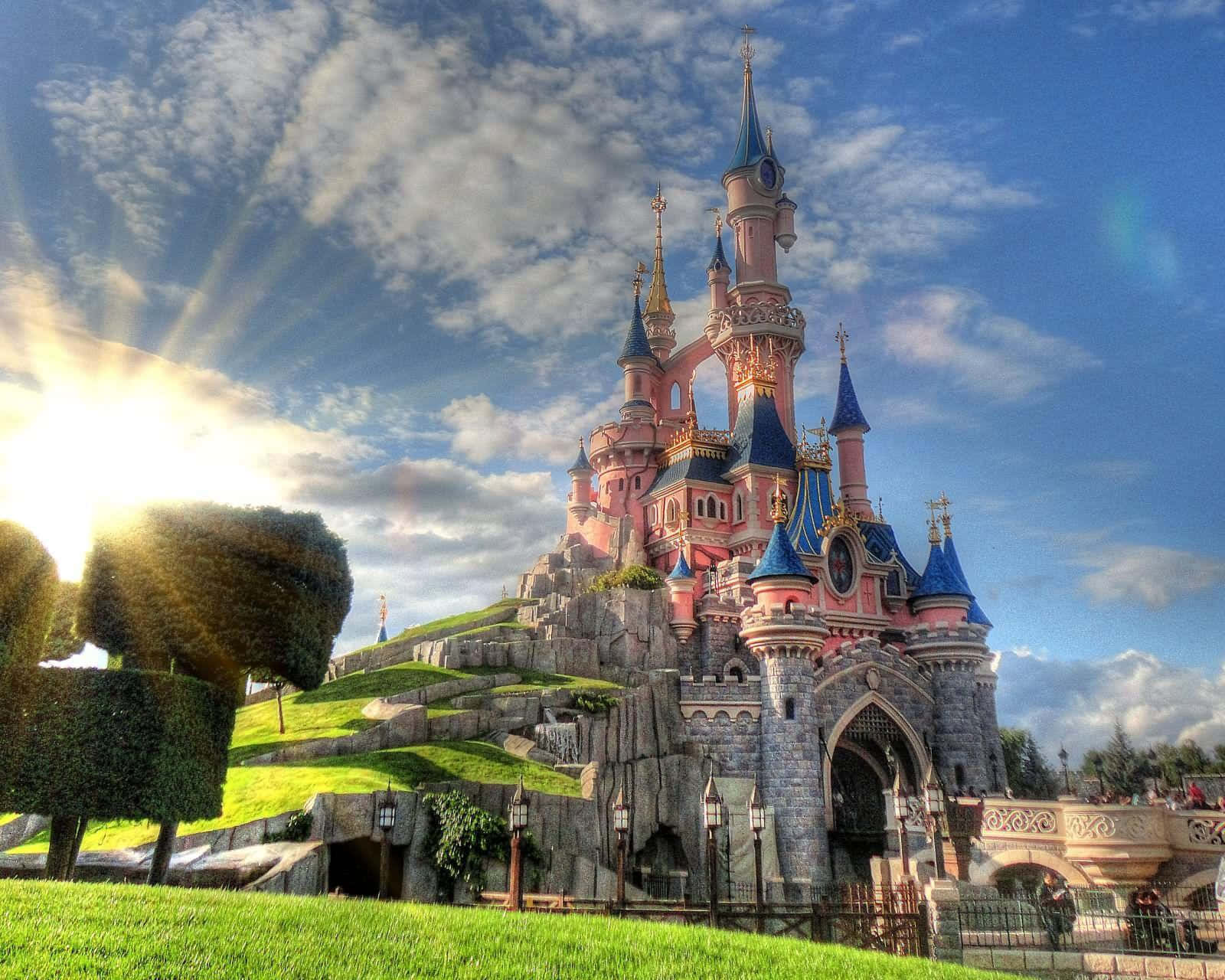 Disneyland Castle 桌布 壁紙 — 一座宏偉的粉紅色和藍色城堡，帶有尖塔，矗立在長滿青草的山丘上，天空明亮，陽光明媚，雲朵點綴。