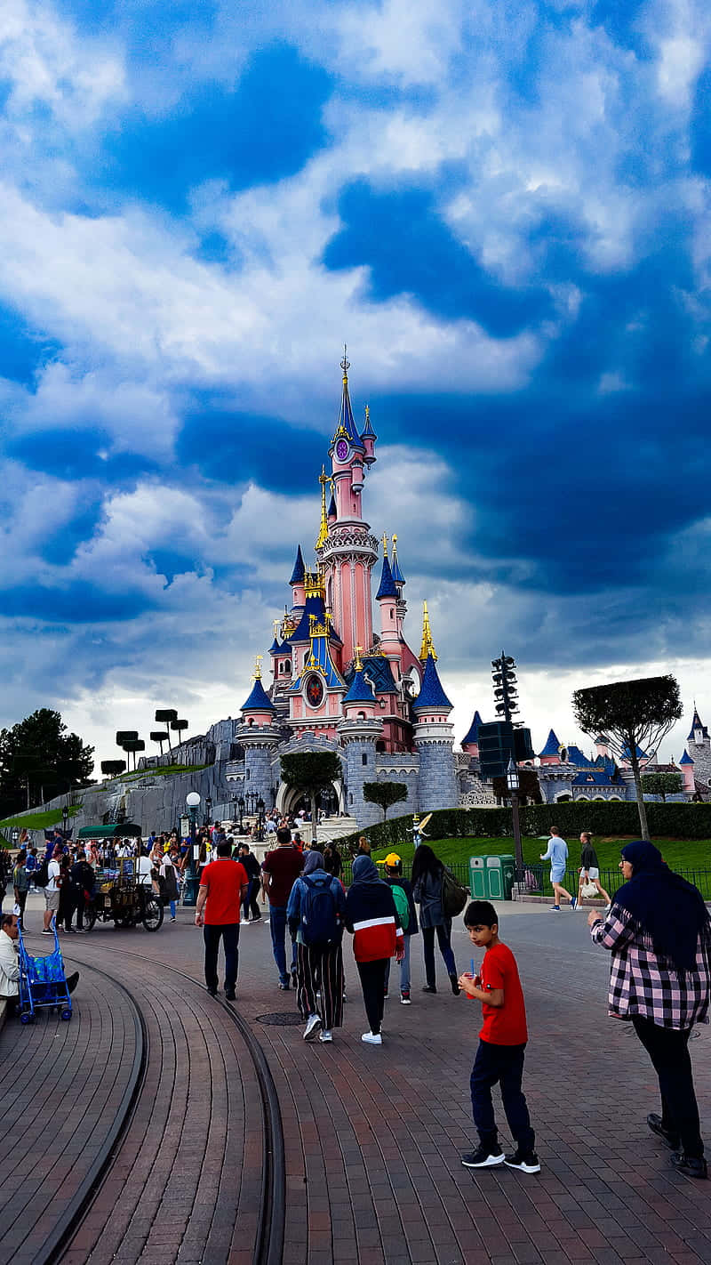 Disneyland 桌布 壁紙 — 一座宏偉的粉紅色和藍色城堡，金色尖塔聳立在戲劇性的多雲天空下，遊客沿著磚砌小徑漫步。