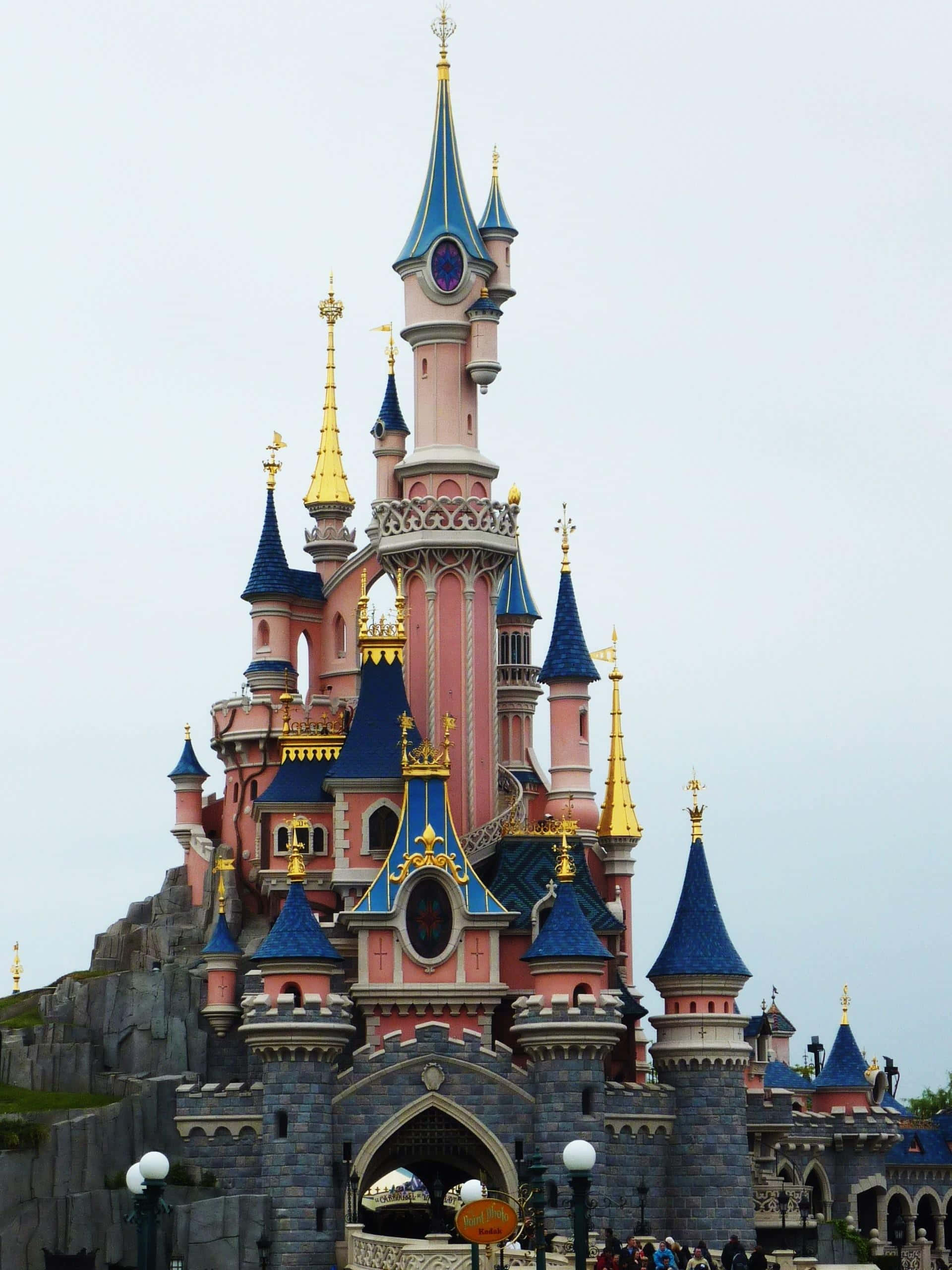 Disneyland 桌布 壁紙 — 一座宏偉的粉紅色和藍色城堡，金色尖塔聳立在多雲的天空下，周圍環繞著石牆和下方的遊客。