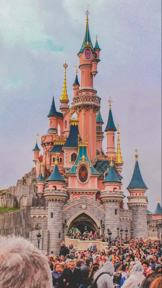 Disneyland Castle 桌布 壁紙 — 一座宏偉的粉紅色和藍色城堡，金色尖塔聳立在熙熙攘攘的人群之上，天空柔和多雲。