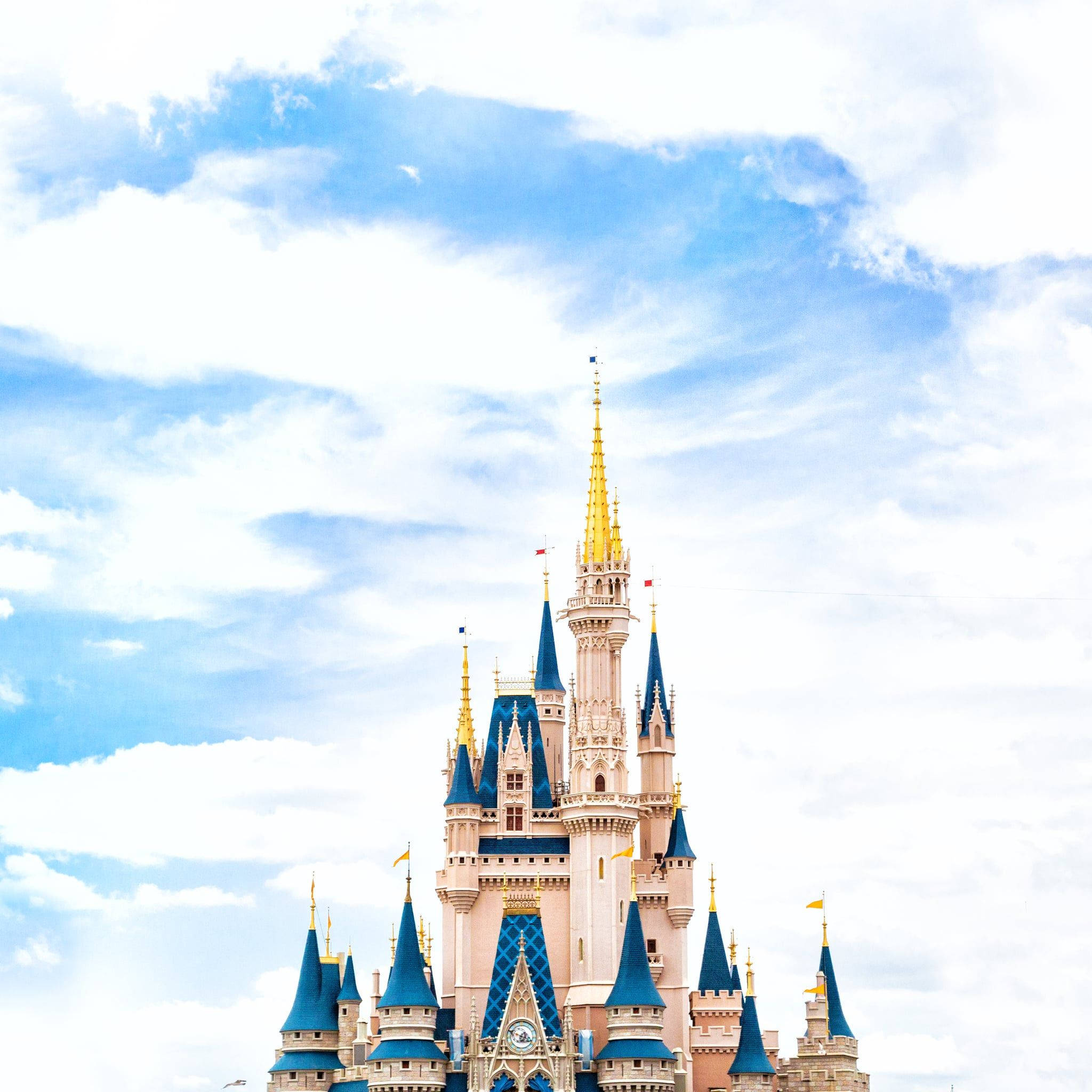 Disney Castle 桌布 壁紙 — 一座宏偉的粉紅色城堡，帶有藍色尖頂和金色點綴，在明亮的藍天和 fluffy 白雲襯托下聳立。