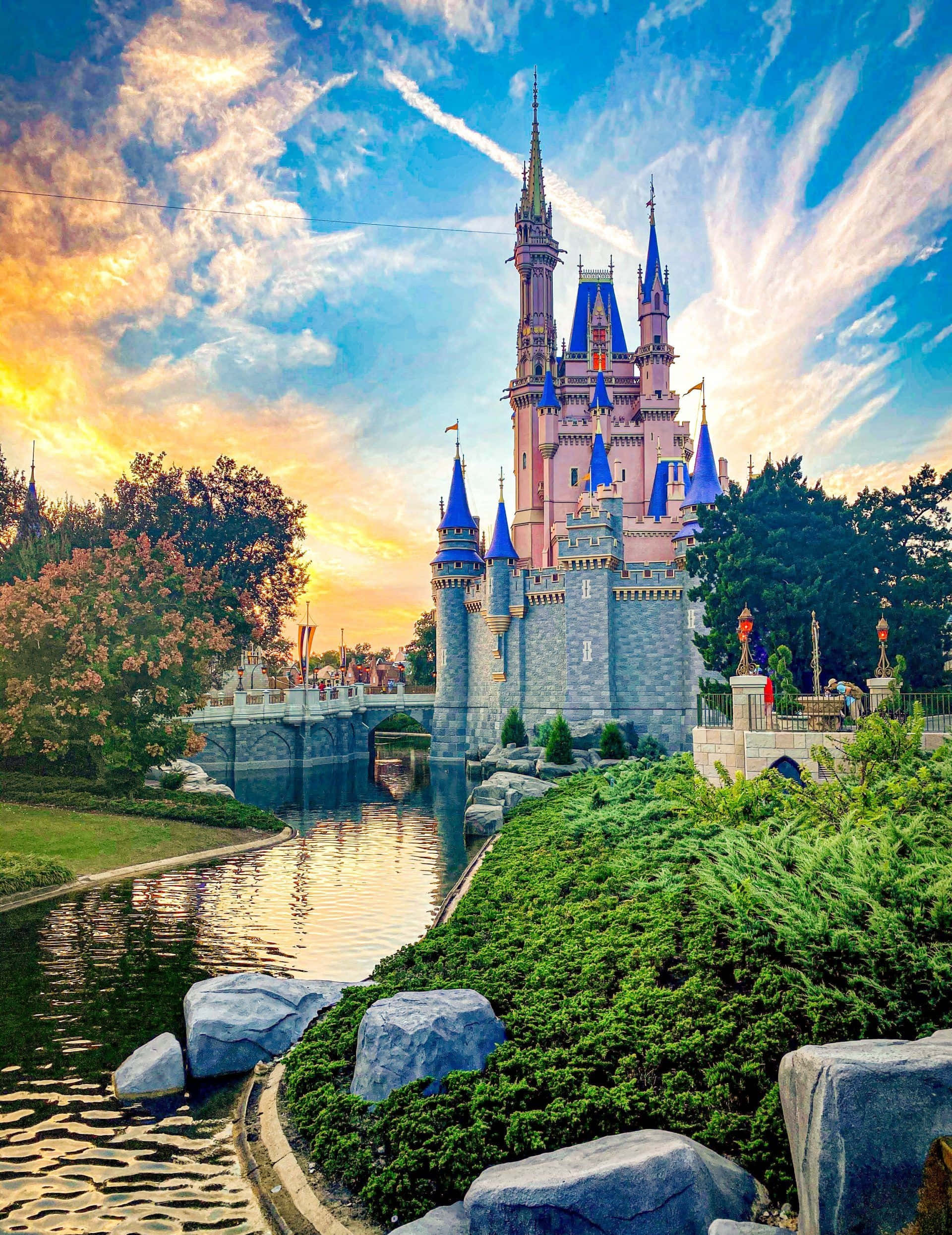 Disney Castle 桌布 壁紙 — 一座宏偉的粉紅色城堡，帶有藍色尖頂，在金色日落時分聳立於寧靜的運河之上，周圍環繞著茂密的綠植和秋樹。