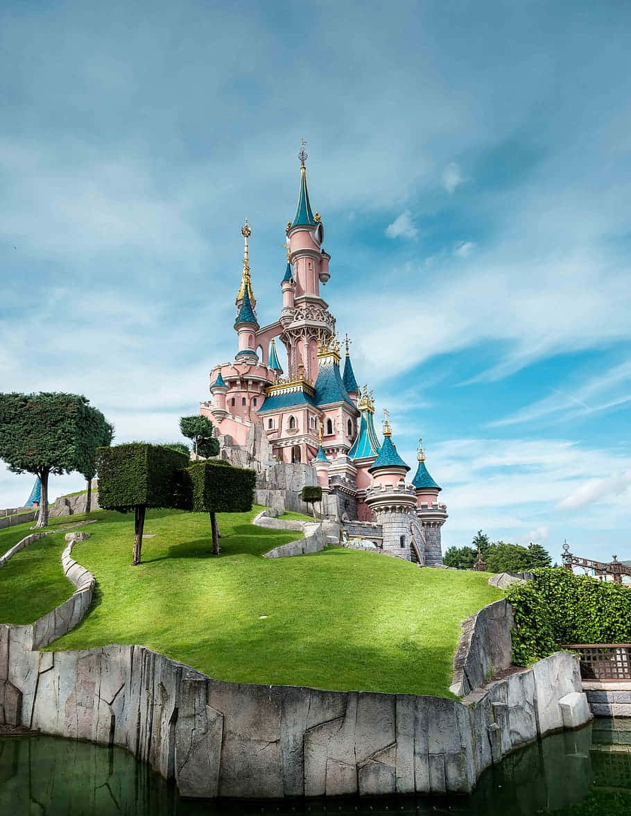 Disneyland Castle 桌布 壁紙 — 一座宏偉的粉紅色城堡，帶有藍色尖頂，聳立在鬱鬱蔥蔥的綠色山丘上，天空湛藍。