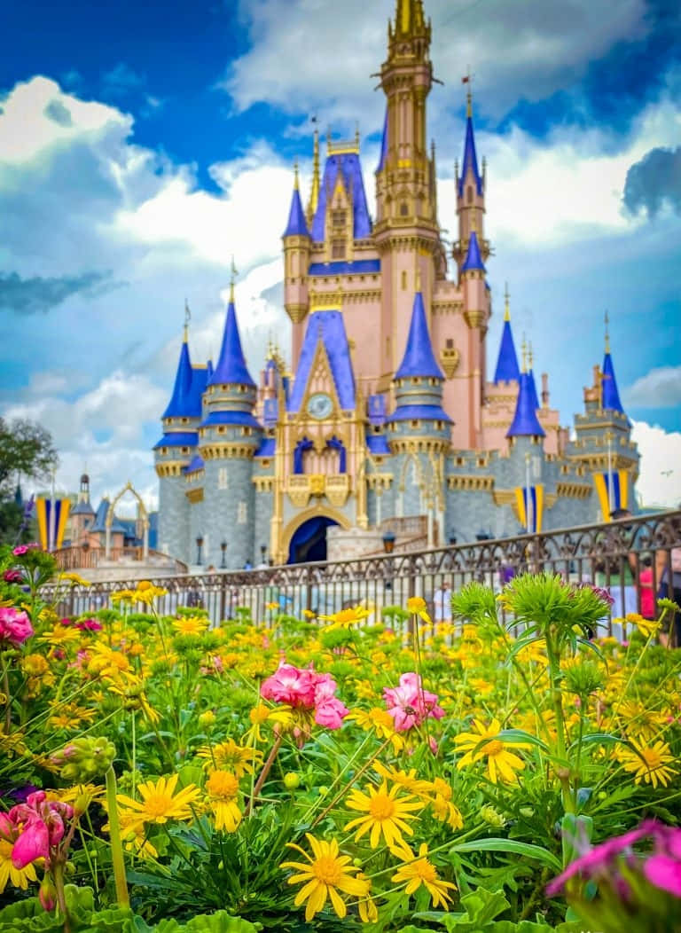 Disney Castle 桌布 壁紙 — 一座宏偉的粉紅色城堡，帶有藍色尖頂，在明亮的藍天下，從一片充滿黃色和粉紅色花朵的鮮豔田野後方升起。