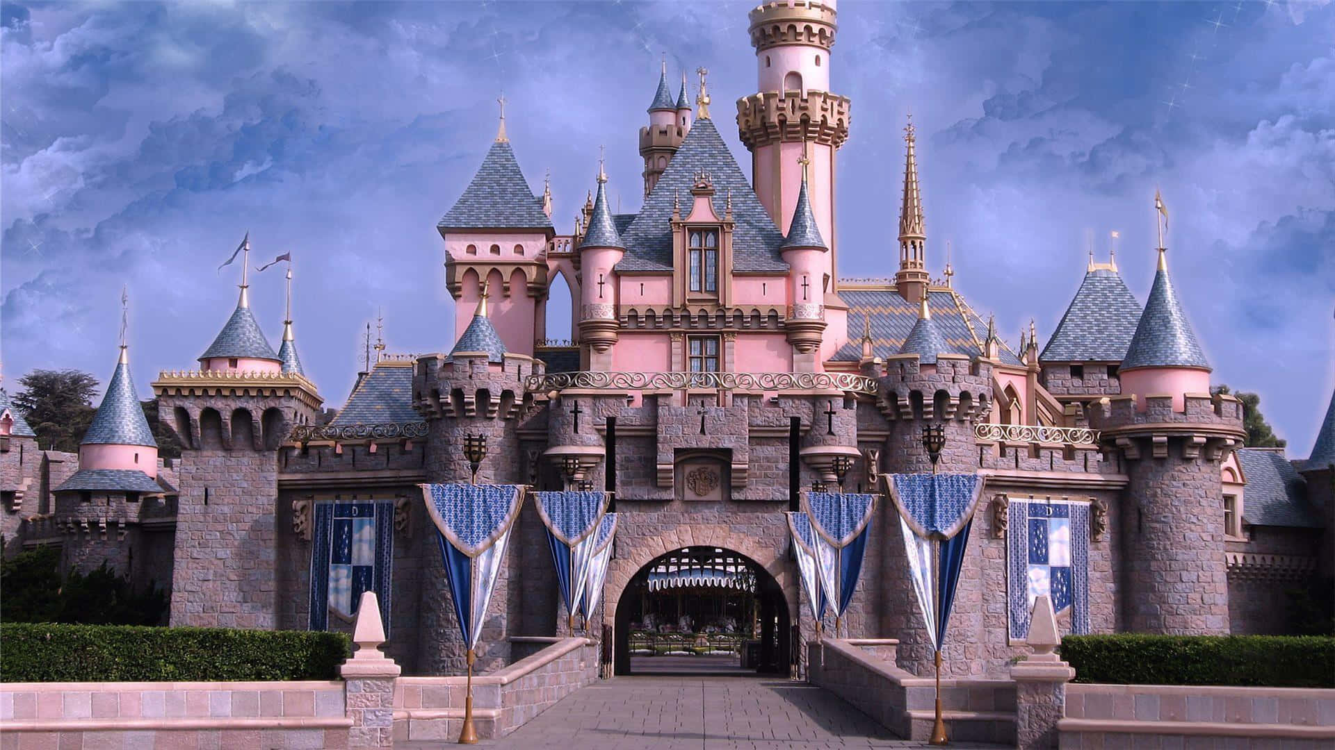 Disneyland 桌布 壁紙 — 一座宏偉的粉紅色城堡，擁有藍色屋頂和金色尖塔，矗立在戲劇性的多雲天空下。