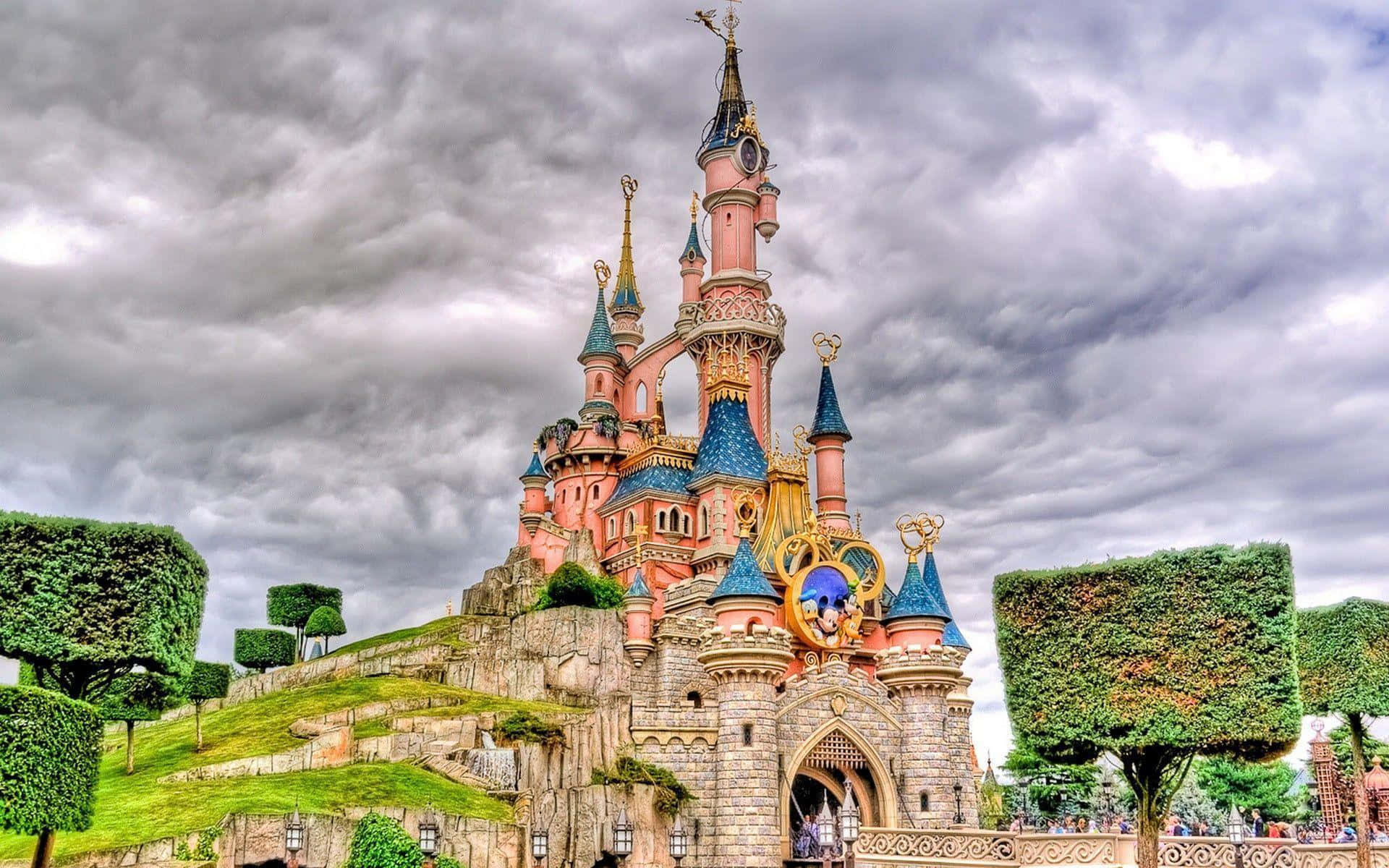 Disneyland 桌布 壁紙 — 一座宏偉的粉紅色城堡，擁有藍色屋頂和金色點綴，矗立在戲劇性的多雲天空下，周圍環繞著修剪整齊的綠色樹籬。